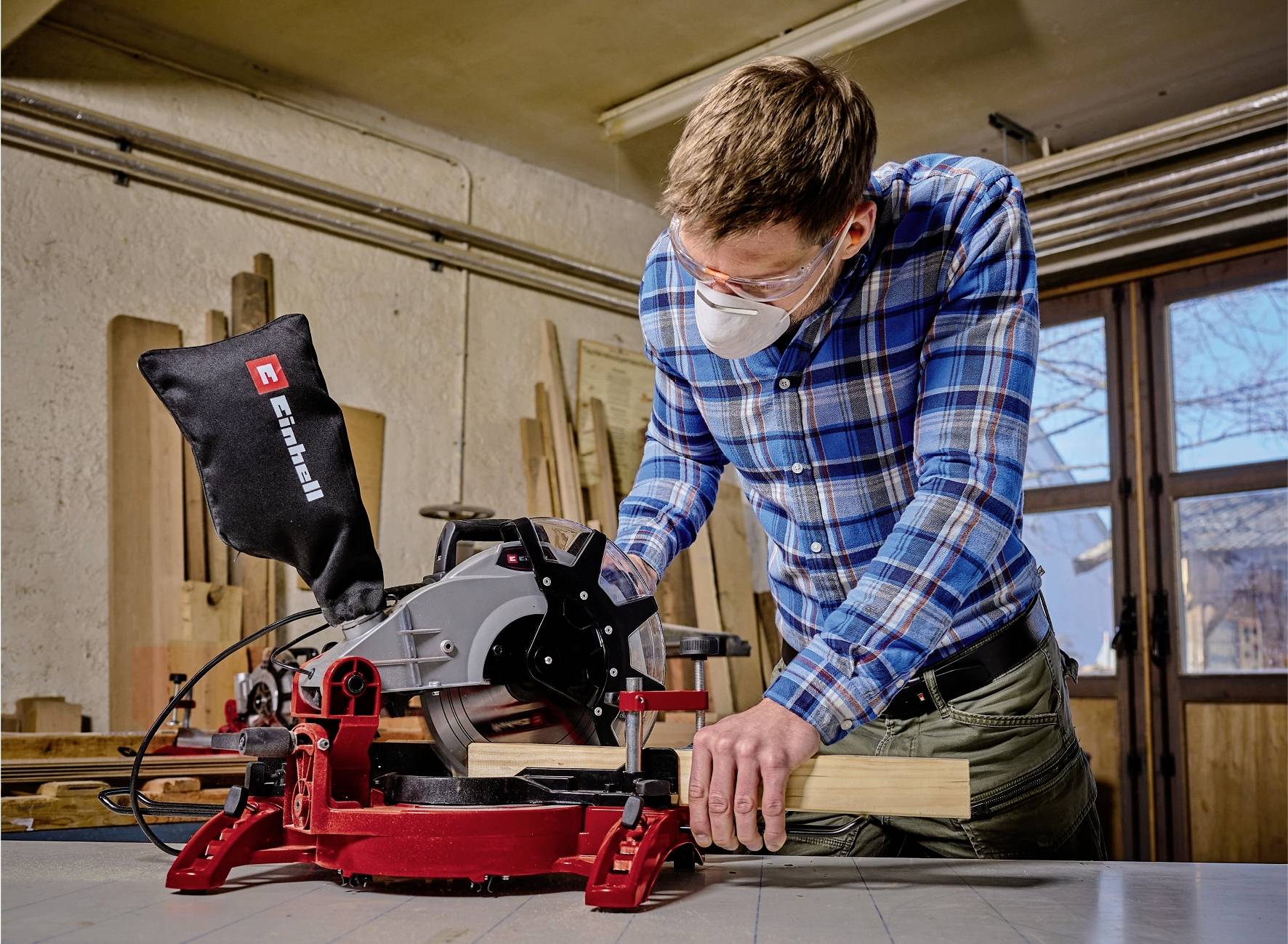 Un homme portant des lunettes et un masque travaille un morceau de bois à l'aide d'une machine rouge et noire dans un atelier. Des fenêtres sont visibles en arrière-plan.