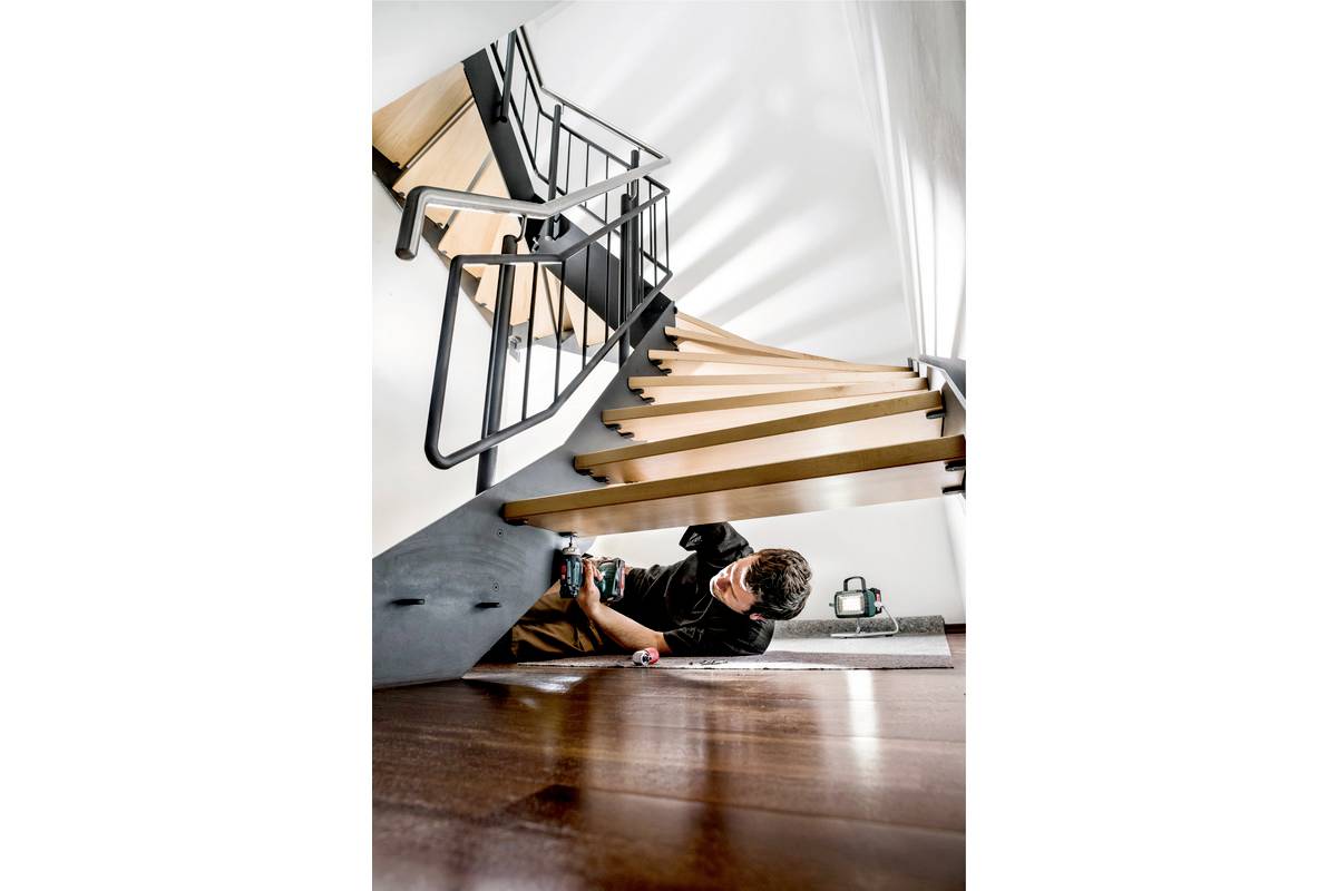 Une personne travaille avec un outil sous un escalier moderne en bois et en métal. Un mur blanc vide se trouve en arrière-plan.