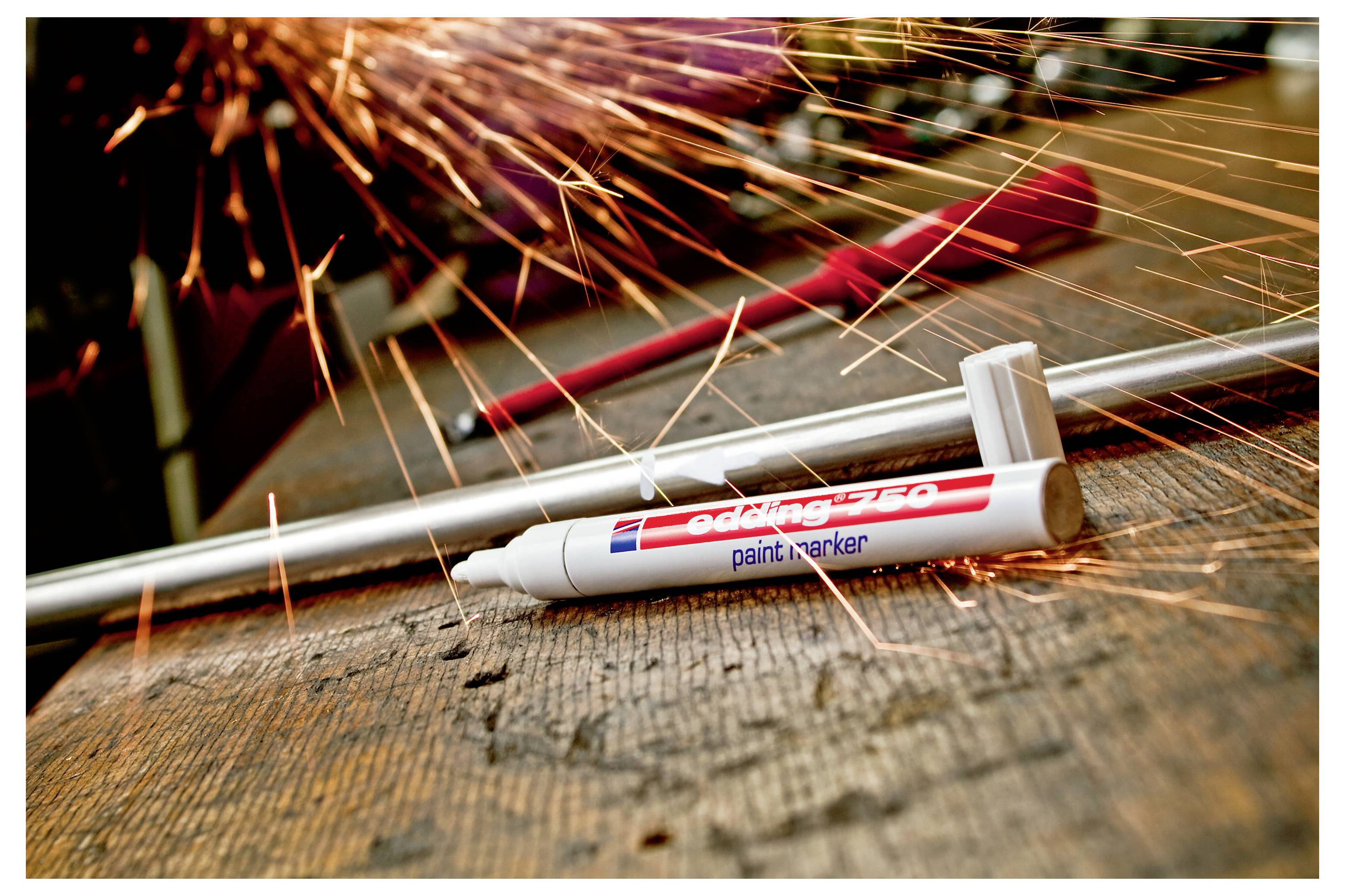 Marqueur de peinture Edding blanc sur une surface en bois usée avec des étincelles oranges qui volent en arrière-plan, suggérant un meulage de métal ou un soudage à proximité.