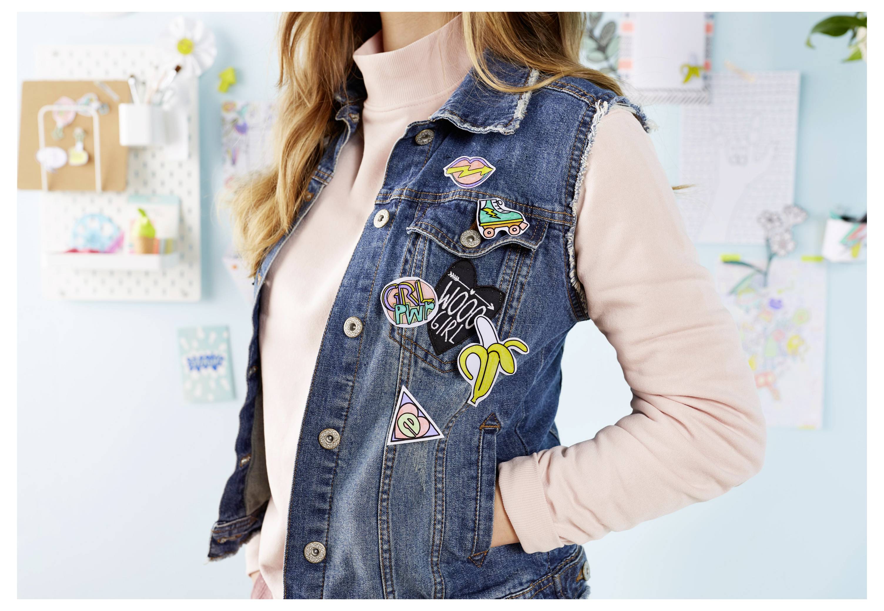 Femme portant un gilet en denim avec des patchs brodés colorés sur un haut rose, photographiée dans une pièce lumineuse avec de l'art mural en arrière-plan.