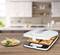 Un sandwich-maker sur une planche en bois, avec deux sandwichs grillés à l'intérieur de l'appareil et une assiette avec deux sandwichs placée à gauche devant.
