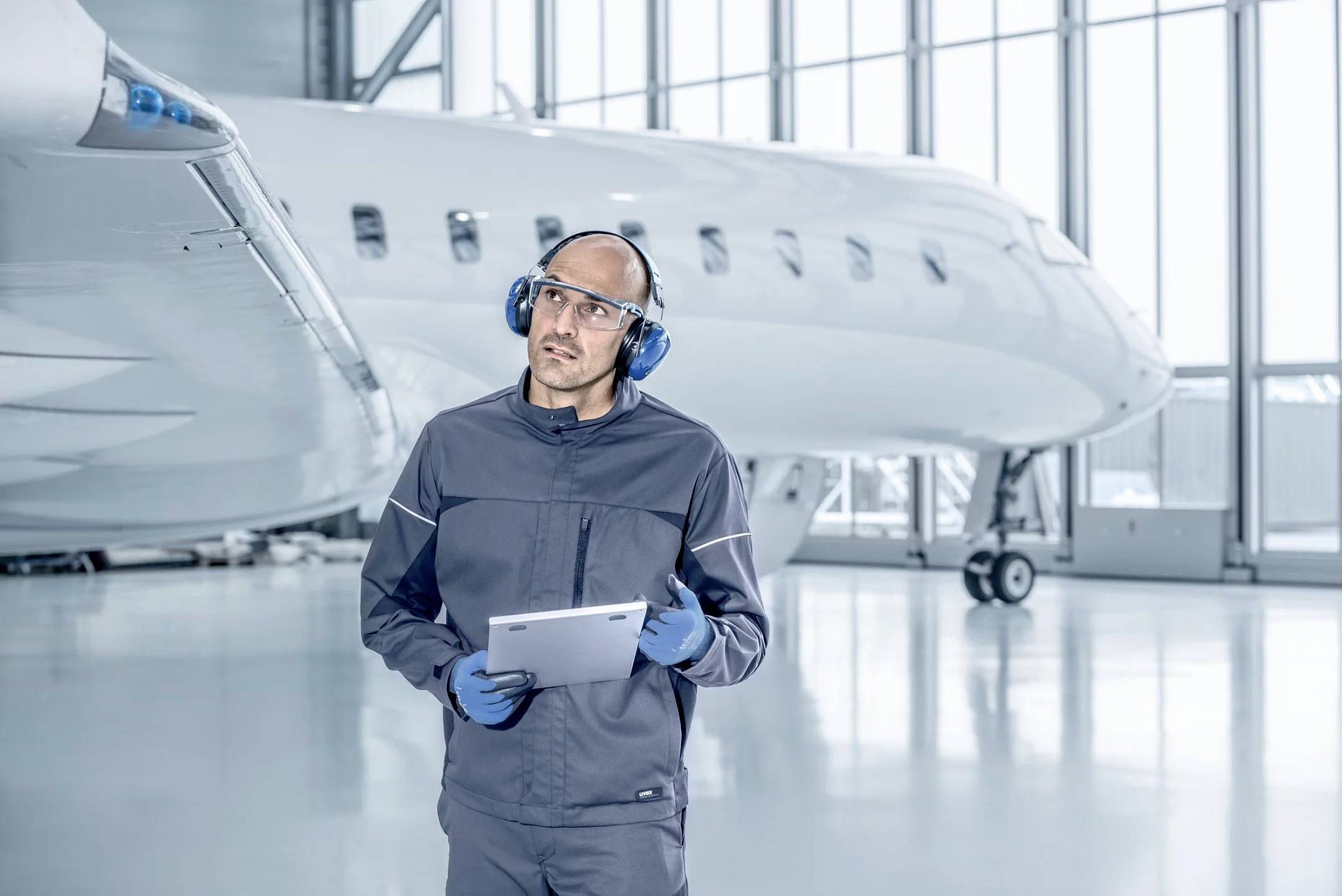 Un technicien en tenue de protection et avec des écouteurs inspecte un avion à l'aide d'une tablette dans un hangar. L'avion est situé en arrière-plan.