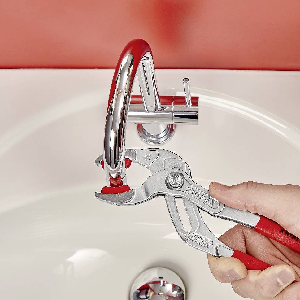 Une personne répare un robinet dans une salle de bain à l'aide d'une pince rouge. Le robinet est installé dans un lavabo.