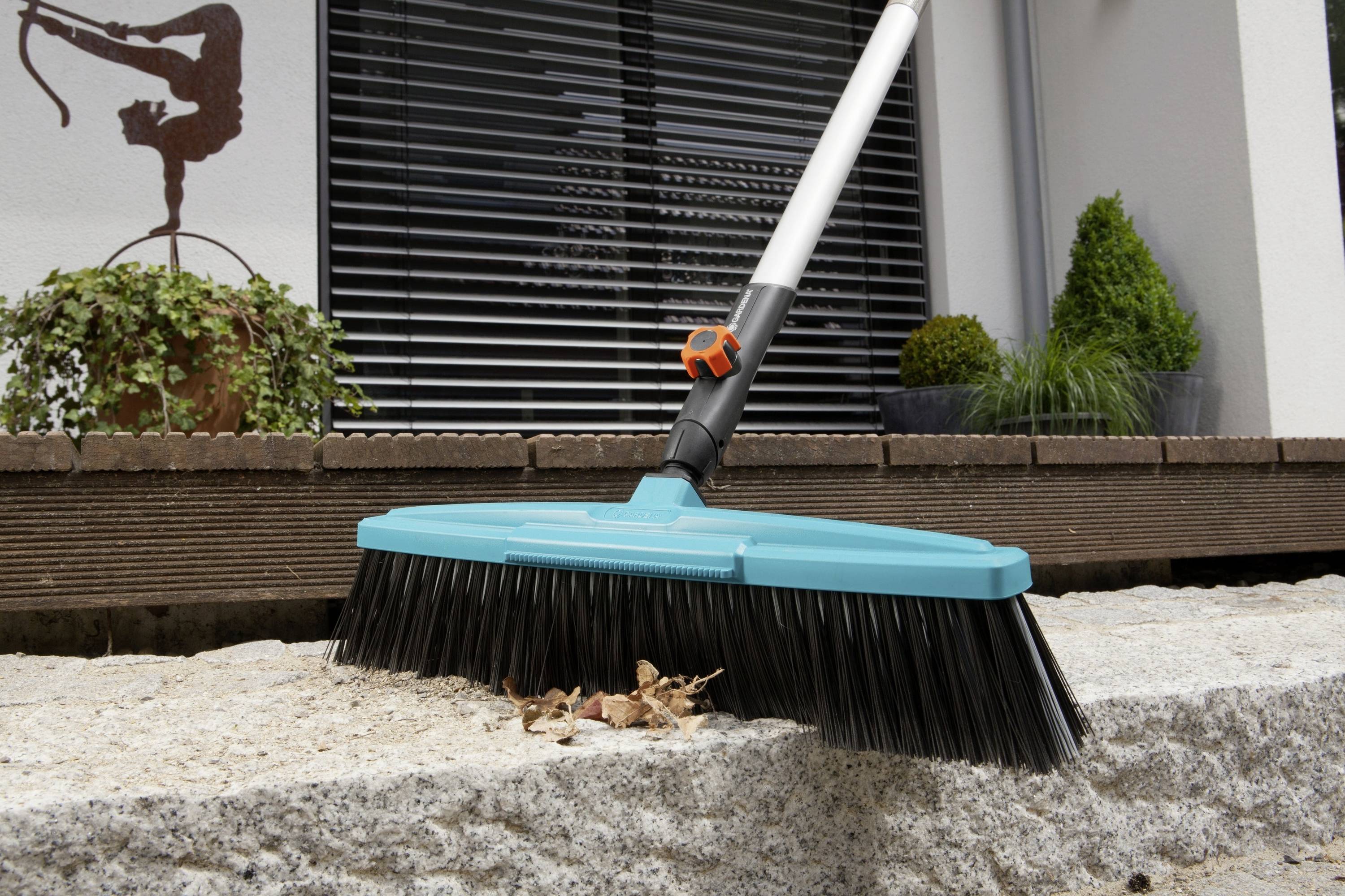 Un balai est posé sur une bordure de pierre avec des feuilles, près d'une maison au design moderne avec des plantes en pot.