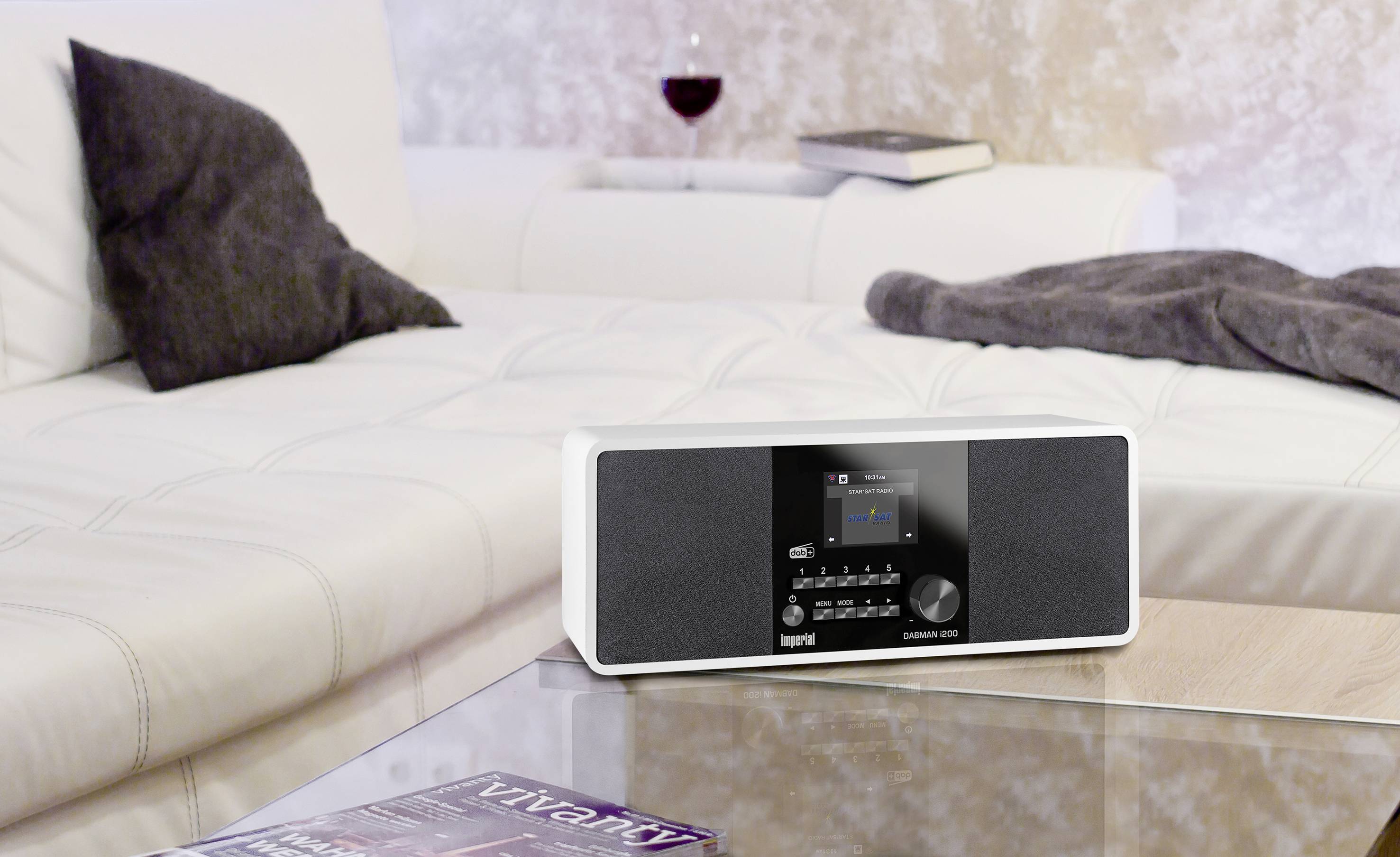 Une radio blanche moderne est posée sur une table en bois dans un salon chaleureux, avec un canapé blanc et un verre de vin en arrière-plan.