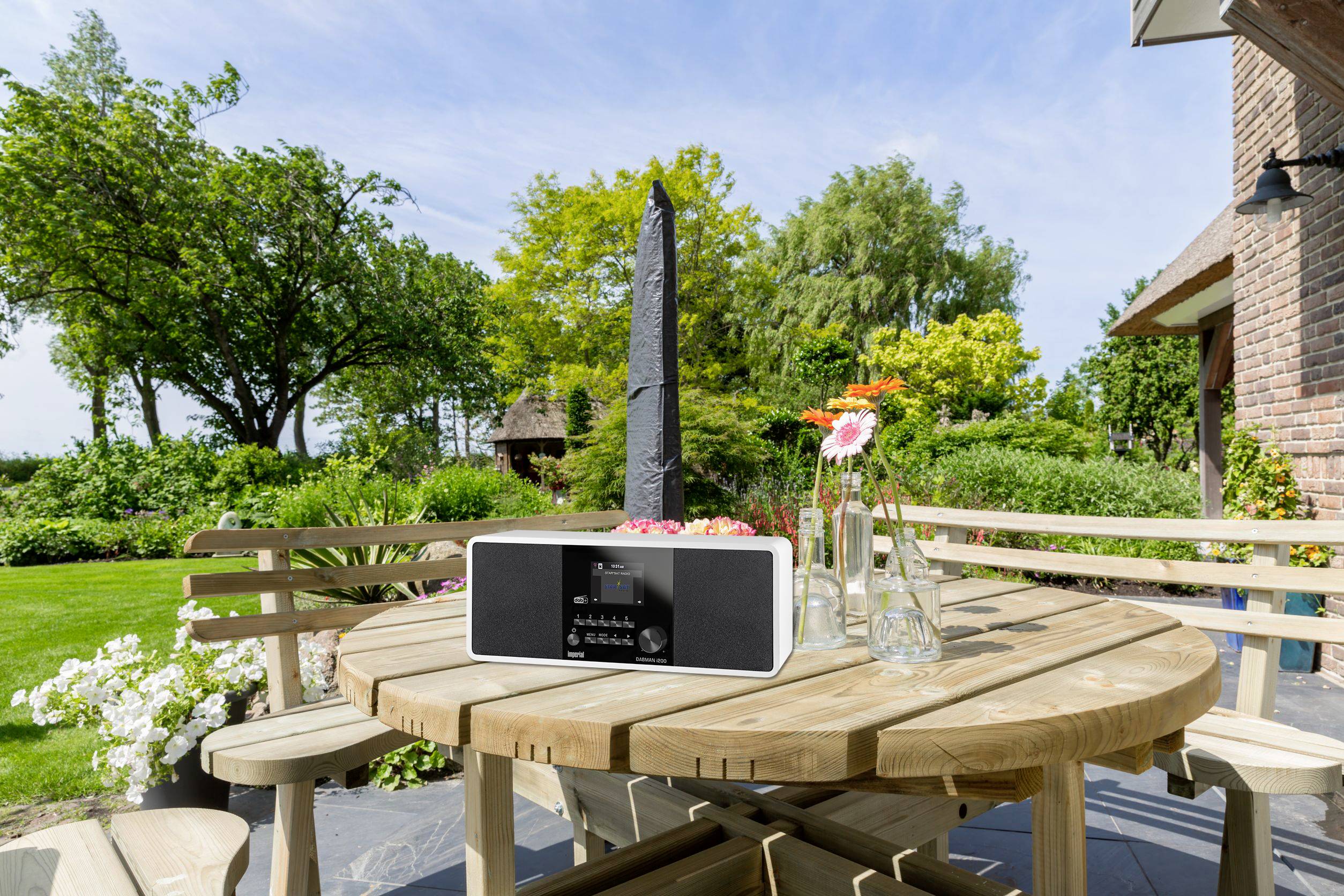 Une radio est posée sur une table ronde en bois dans le jardin. Entourée de fleurs en bloom et d'arbres par une journée ensoleillée.