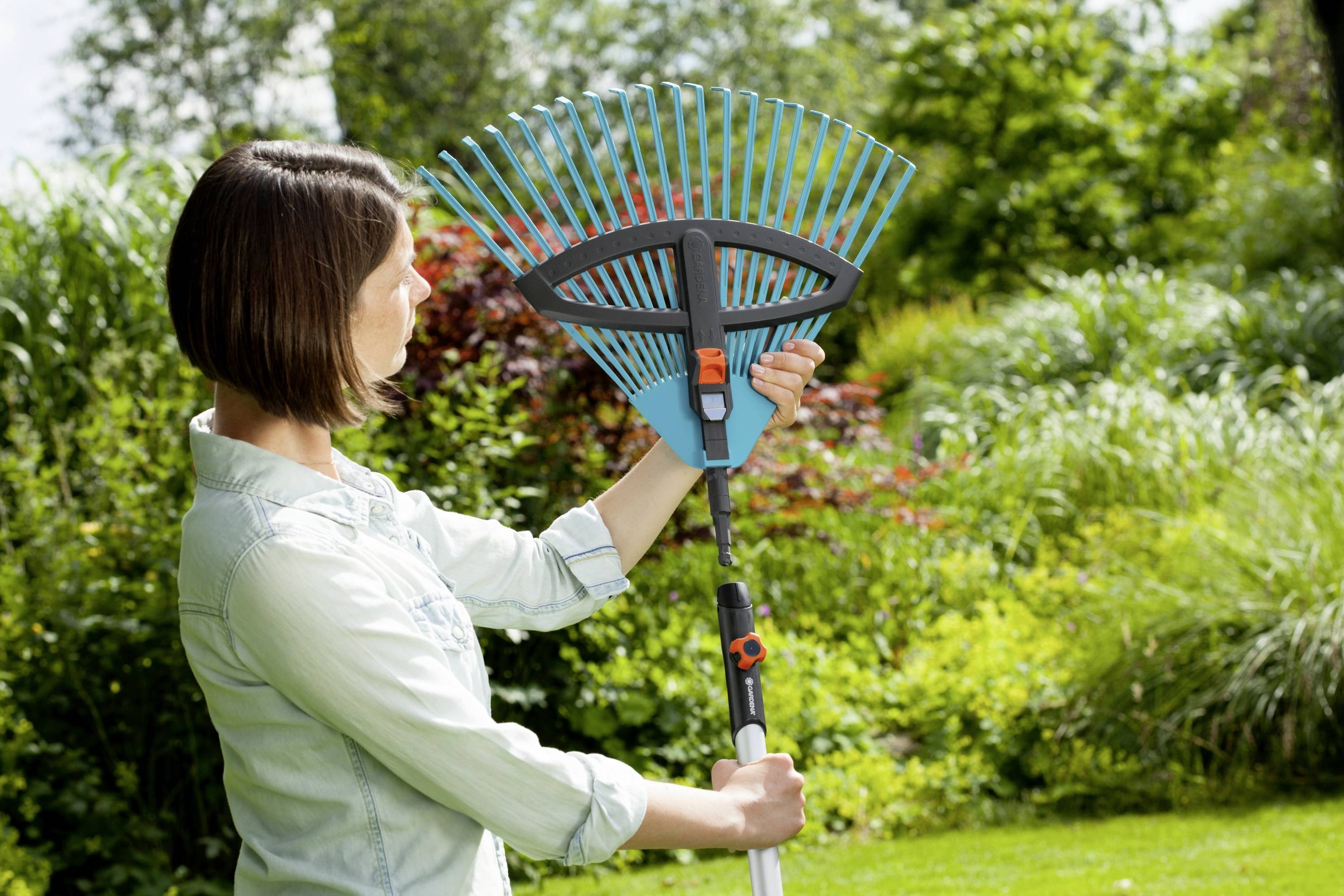 Une personne tient un râteau réglable dans un jardin. Entourée de feuillage vert et de plantes. Elle vérifie le réglage de l'outil.