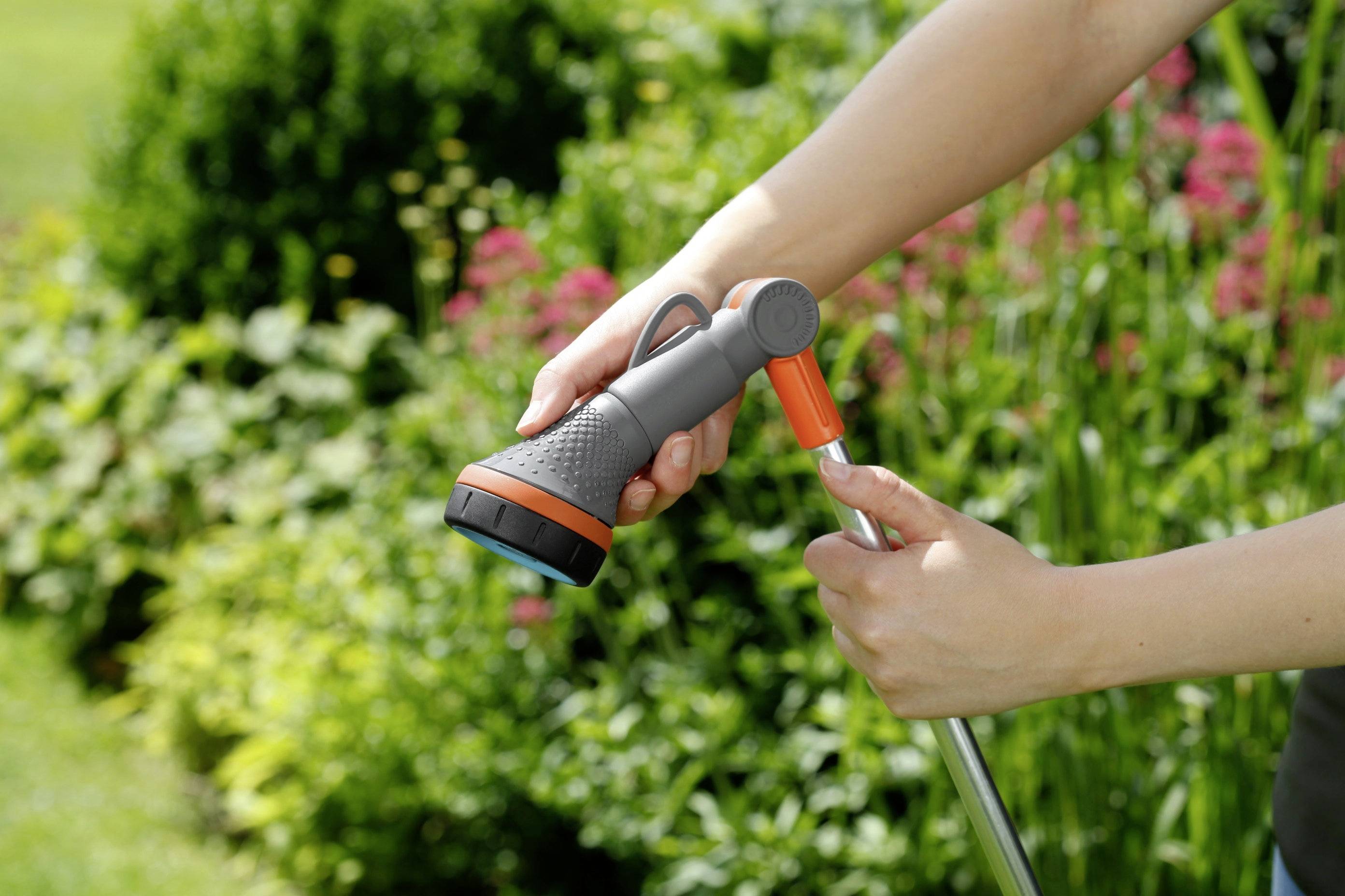 Une personne tient un embout de tuyau d'arrosage gris et orange, entouré de plantes vertes en arrière-plan.