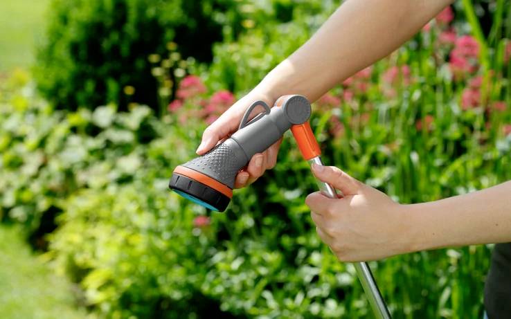 Une personne règle l'embout d'un tuyau d'arrosage avec un mélange de plantes vertes et de fleurs en pleine floraison en arrière-plan.