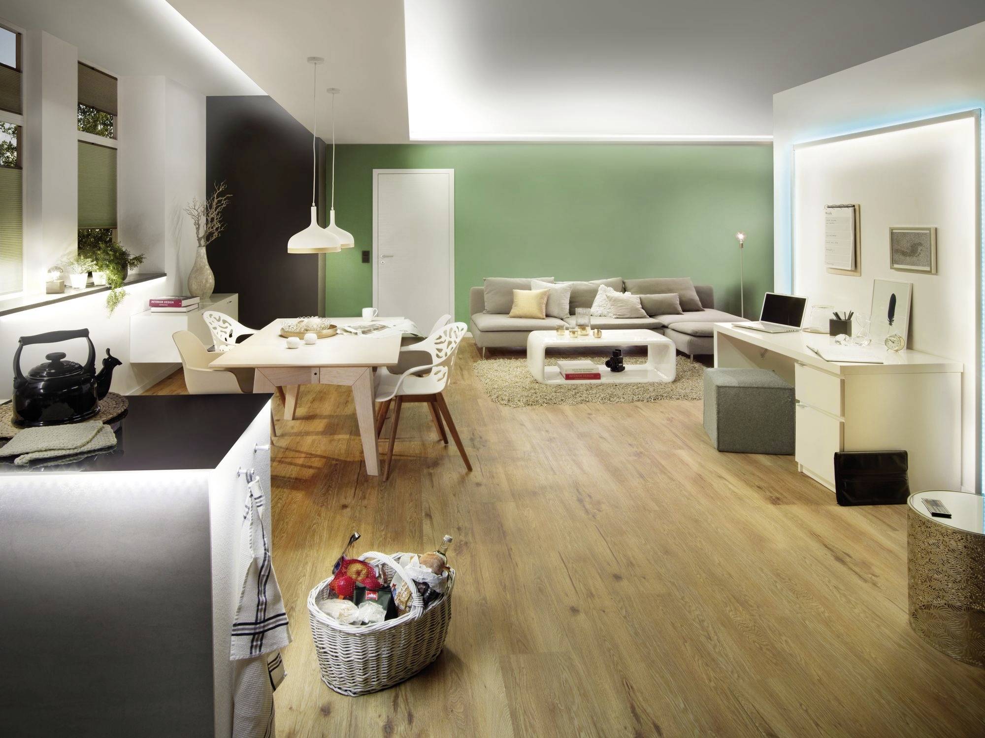 Grand salon moderne avec un sol en bois, un mur vert, une table à manger avec des chaises blanches, un canapé et un bureau. Aménagement lumineux et minimaliste.