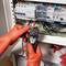 Un électricien portant des gants orange vérifie l'alimentation électrique avec un multimètre dans un armoire électrique ouverte.