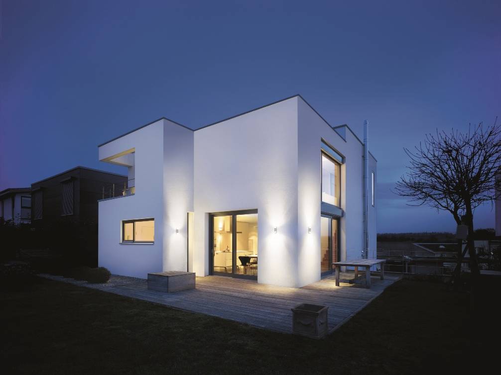 Maison moderne à deux étages avec toit plat, grandes fenêtres et façade éclairée, entourée d'un jardin au crépuscule.