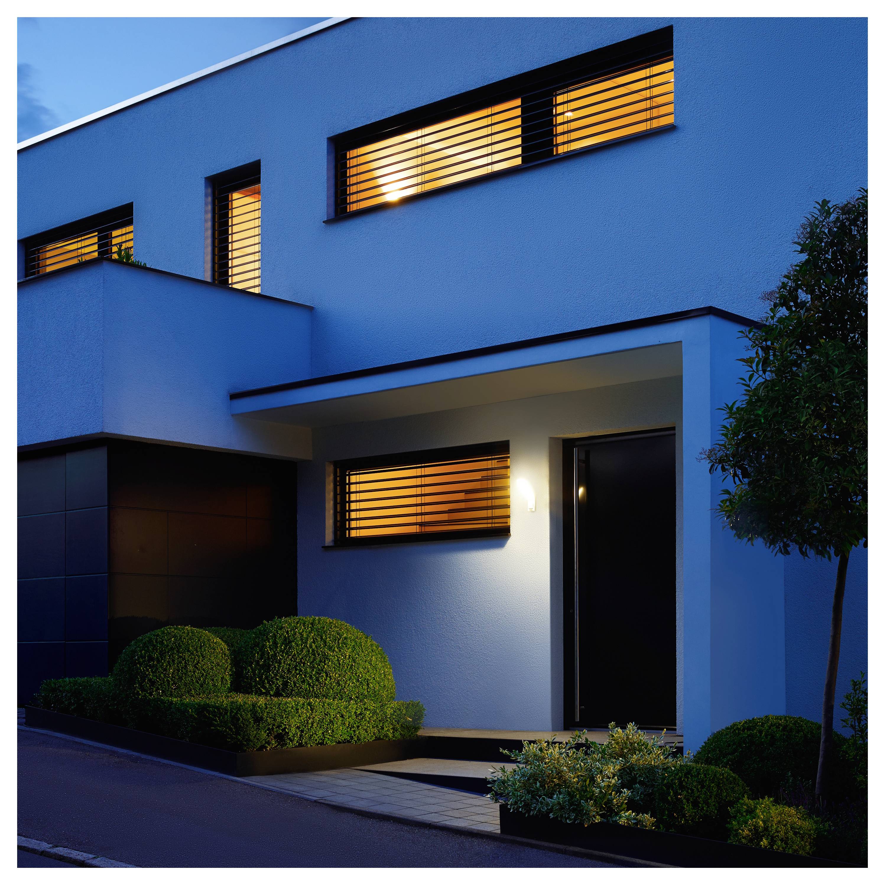 Extérieur moderne d'une maison à deux étages au crépuscule avec des fenêtres illuminées, une lumière de porche avant, des arbustes manucurés et une allée sous un ciel bleu en soirée.