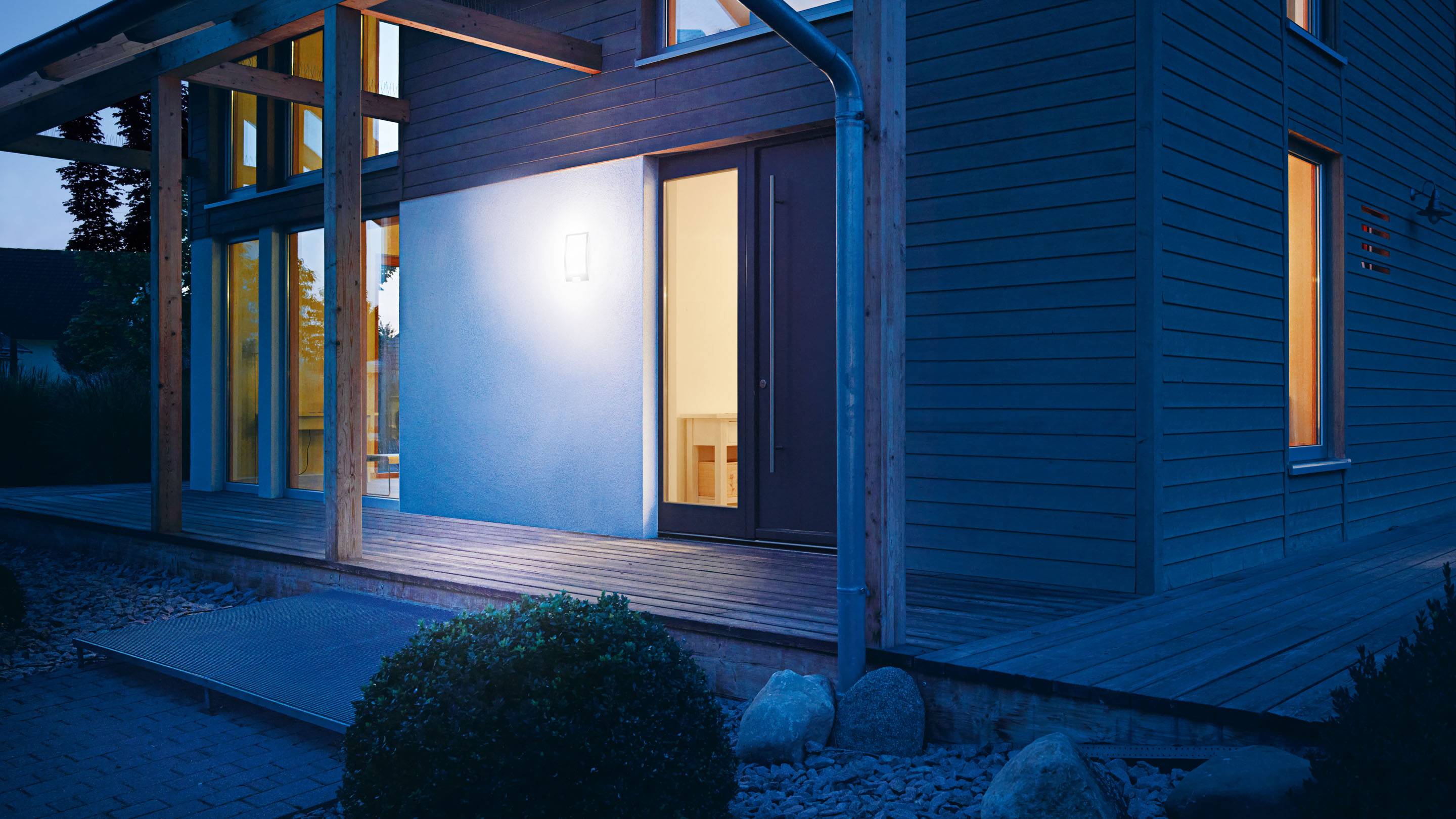 Une maison moderne en bois, illuminée la nuit, avec de grandes fenêtres et une véranda couverte, entourée de plantes et de pierres.