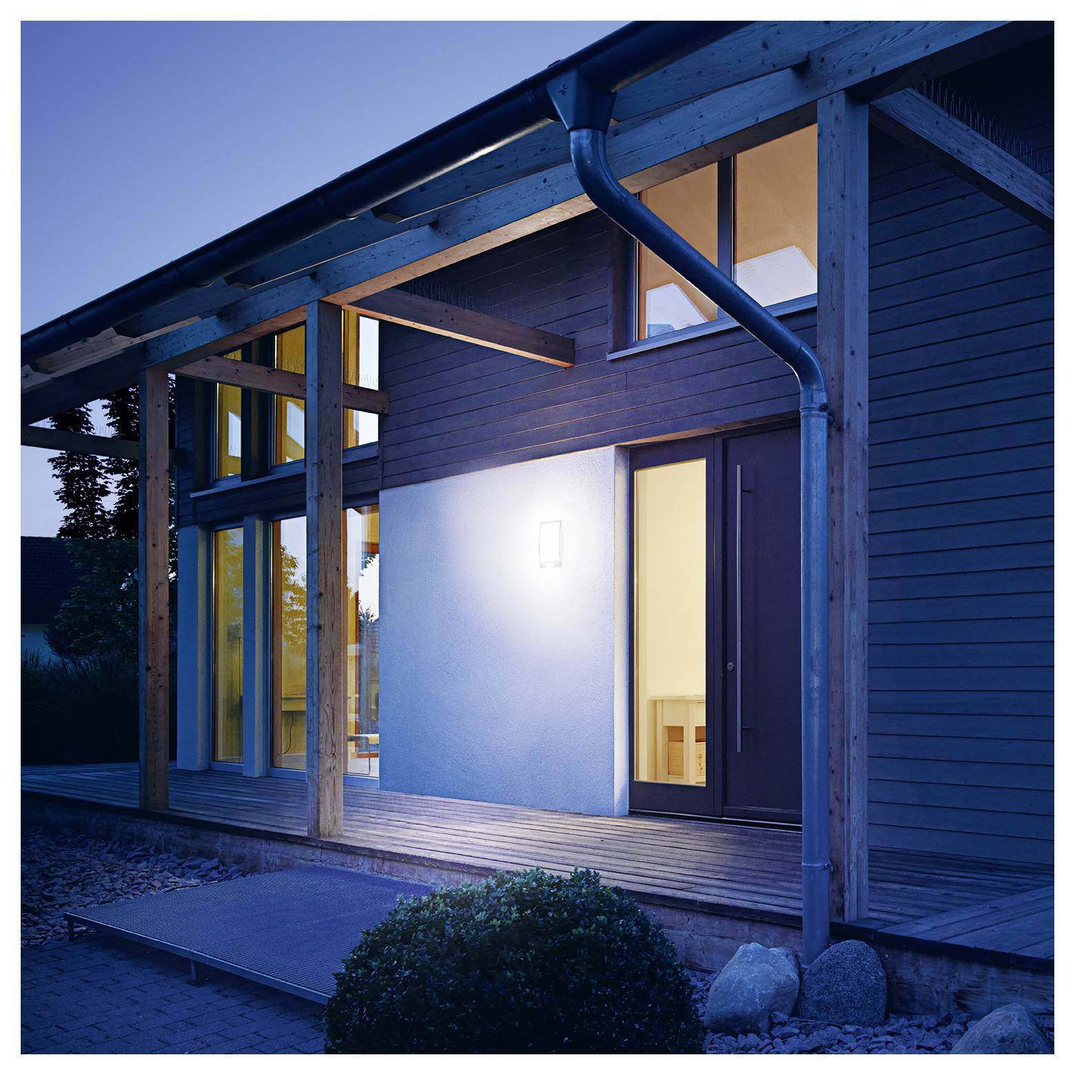 Extérieur d'une maison moderne au crépuscule avec un revêtement en bois, de grandes fenêtres, un porche couvert, et une porte illuminée qui brille de l'intérieur.