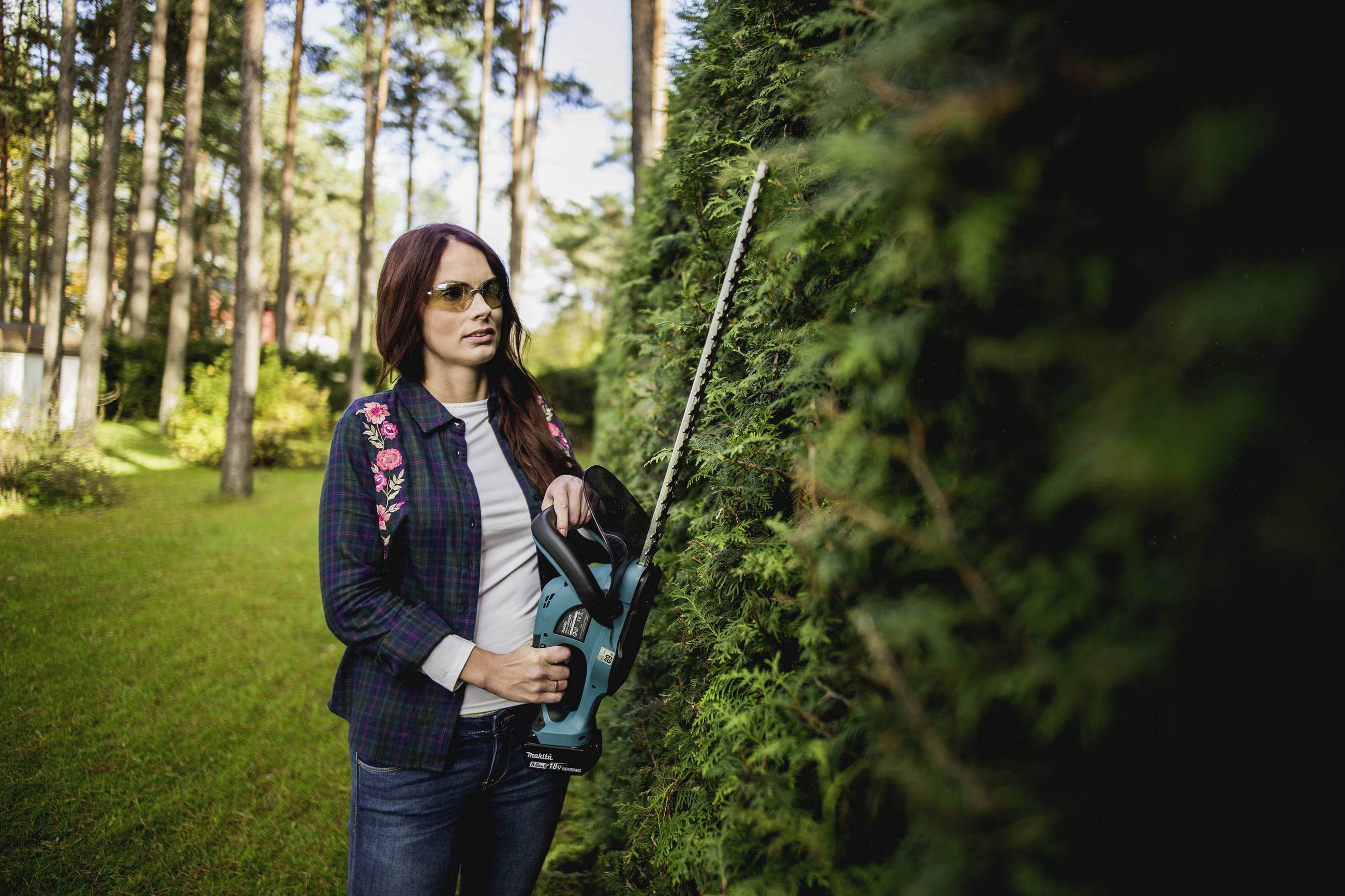 Femme portant des lunettes de soleil taillant une grande haie dans le jardin avec un taille-haie électrique. Environnement entouré d'arbres et de pelouse.