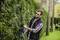 Un homme taille une haie dans un jardin à l'aide d'un taille-haie électrique. Il porte des lunettes de protection et un gilet de sécurité pour se protéger.