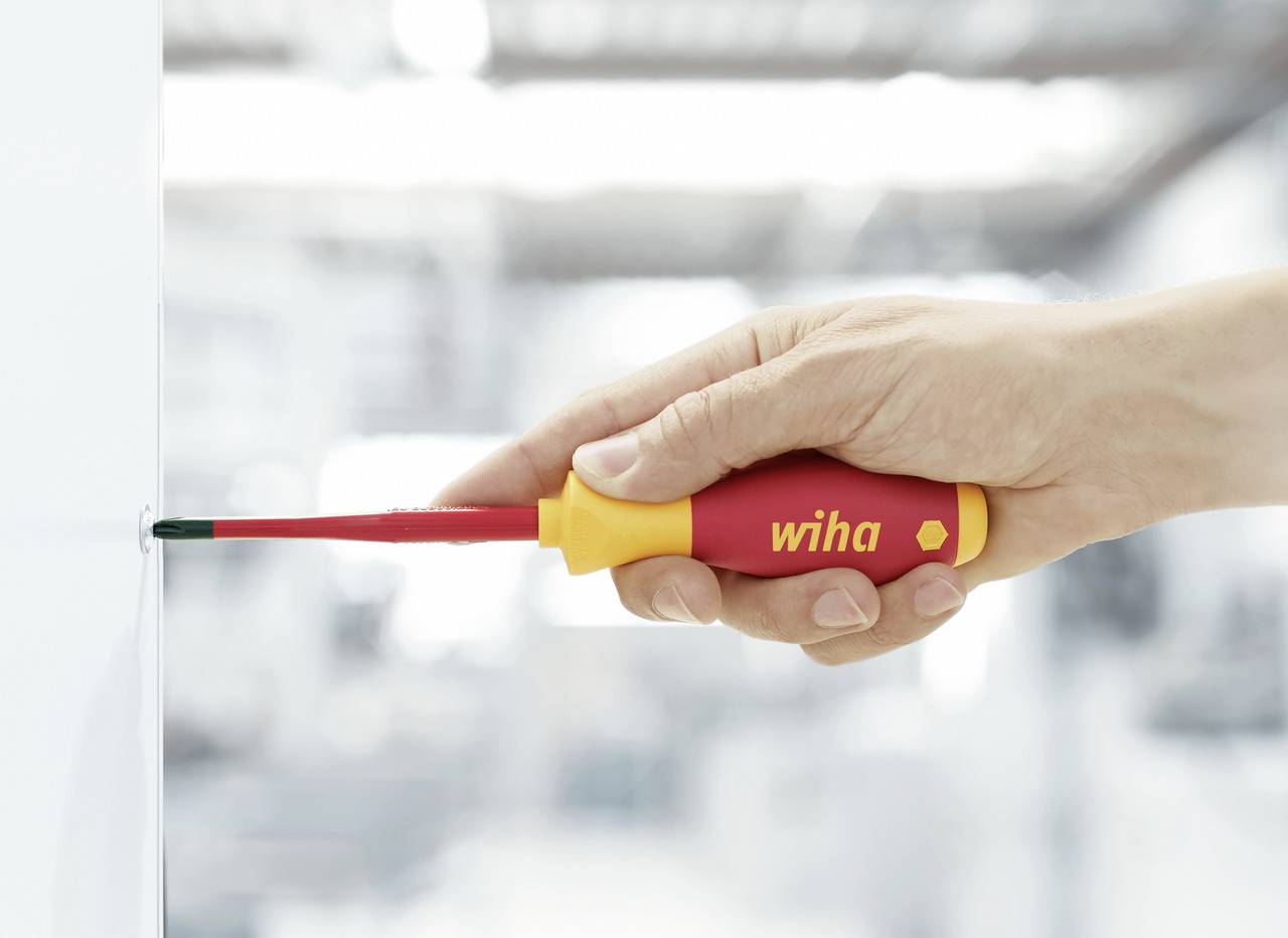 Une main tient un tournevis isolé et visse une vis dans une surface blanche. Le tournevis est rouge et jaune avec le texte 'wiha'.