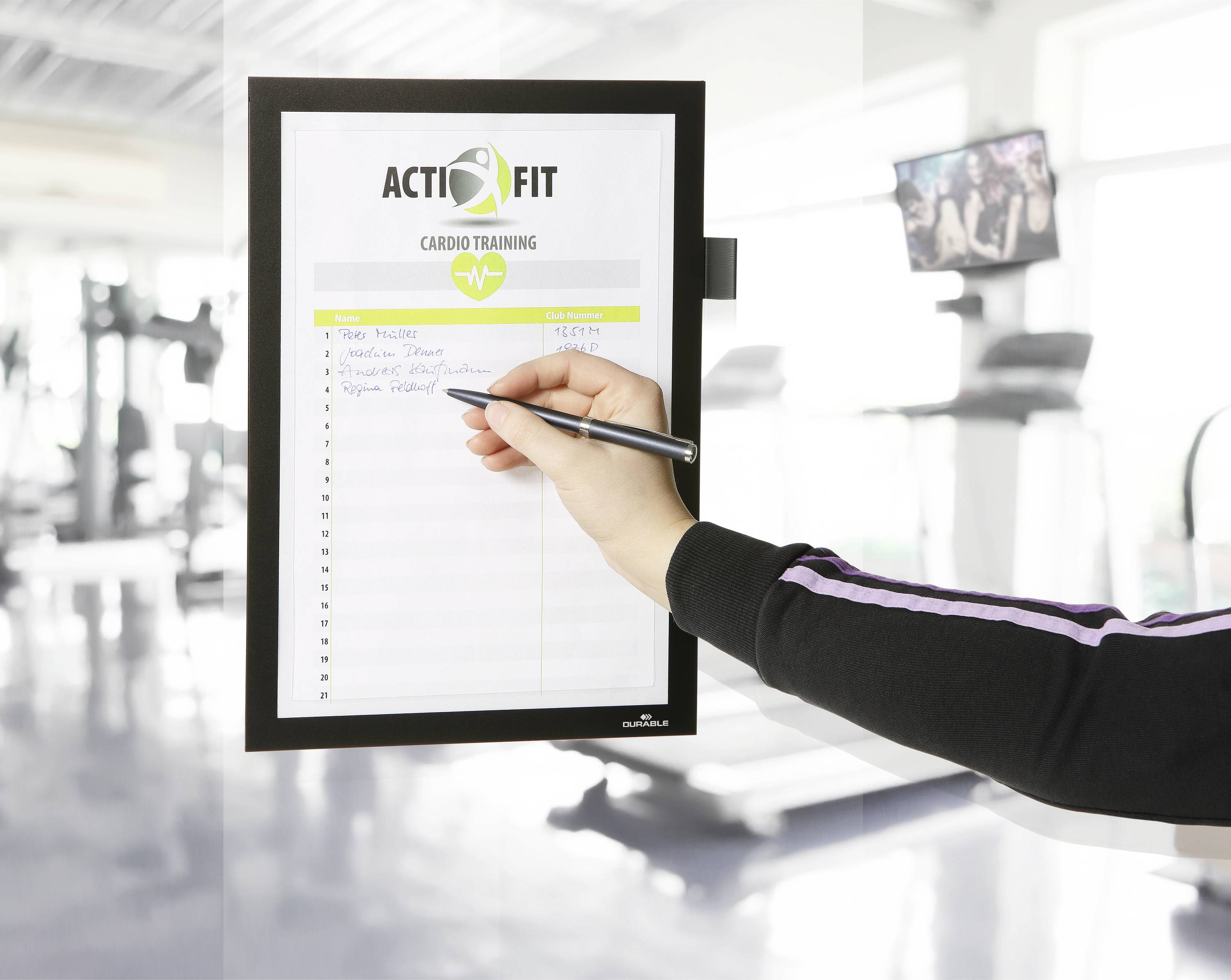 Une personne remplit un formulaire avec un stylo sur un support de presse-papiers dans une salle de fitness, représentant un 'Entraînement Cardio ACTIFIT'.