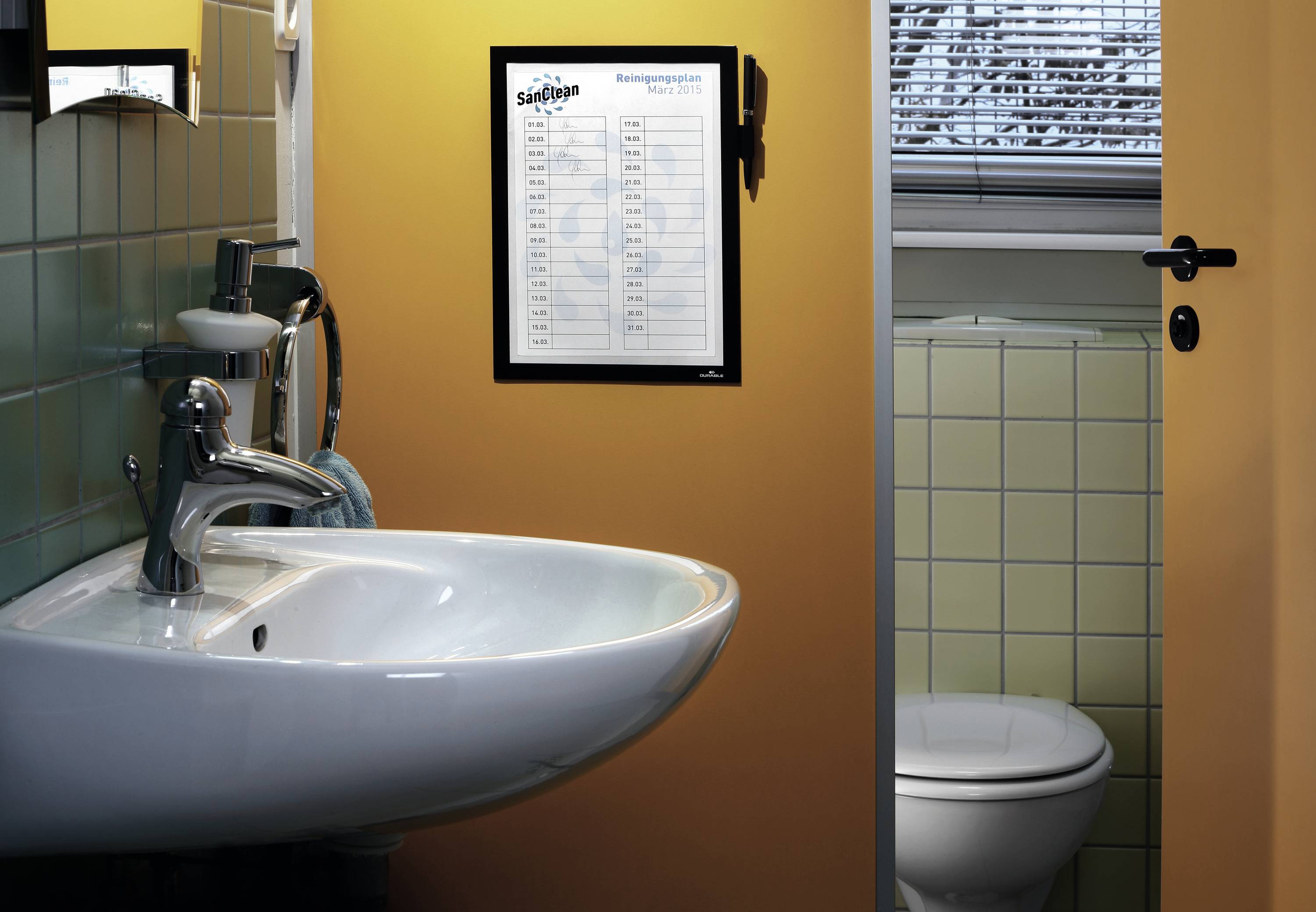 Lavabo et miroir dans une salle de bain orange. Un planning de nettoyage est accroché au mur. Une porte ouverte mène aux toilettes.