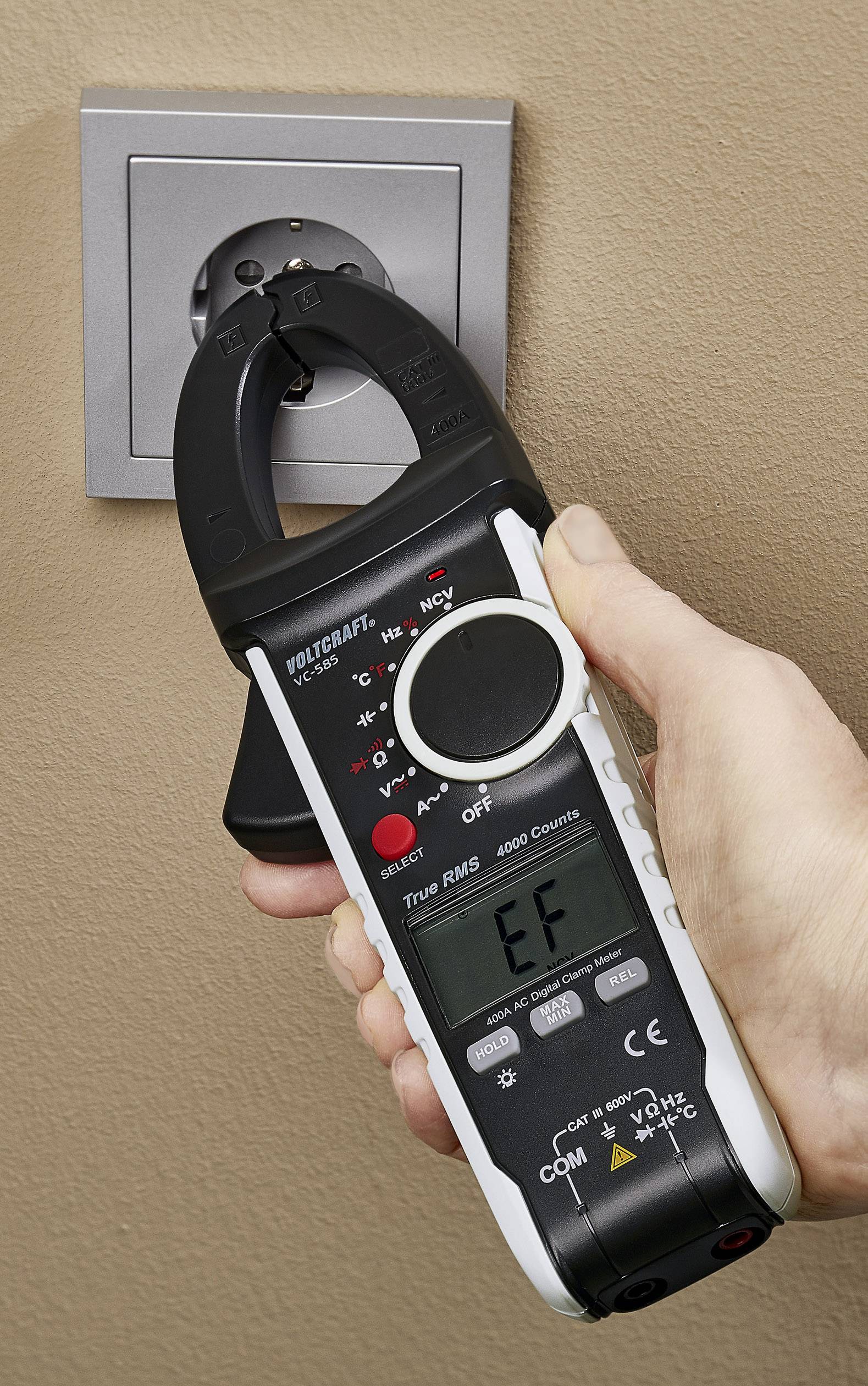 Une main tient un ampèremètre à pince, connecté à une prise électrique pour effectuer des mesures électriques.