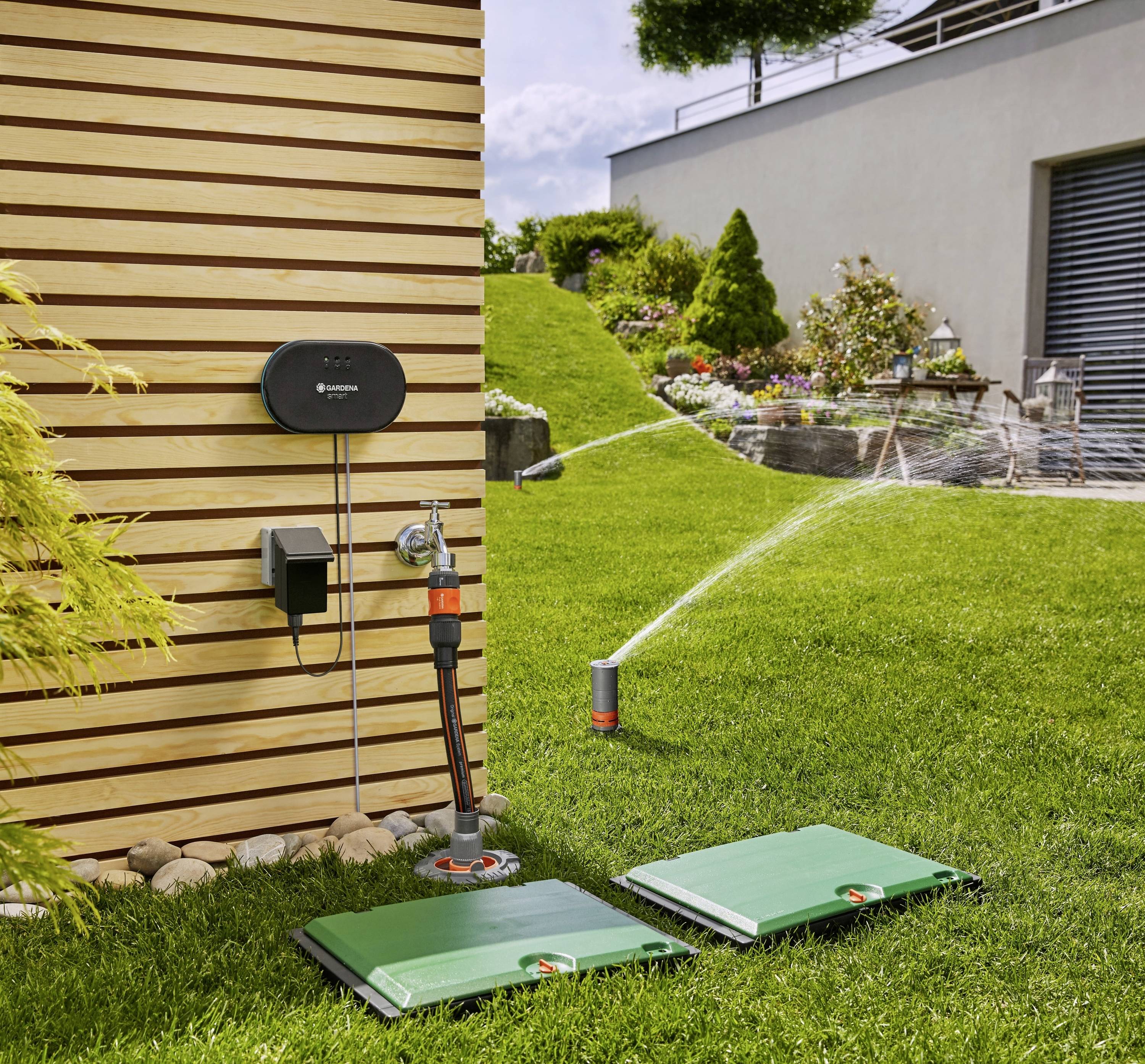Système d'irrigation automatisé dans un jardin avec des sprinklers et une unité de contrôle fixée sur un mur en bardage de bois, pelouse et parterre de fleurs.