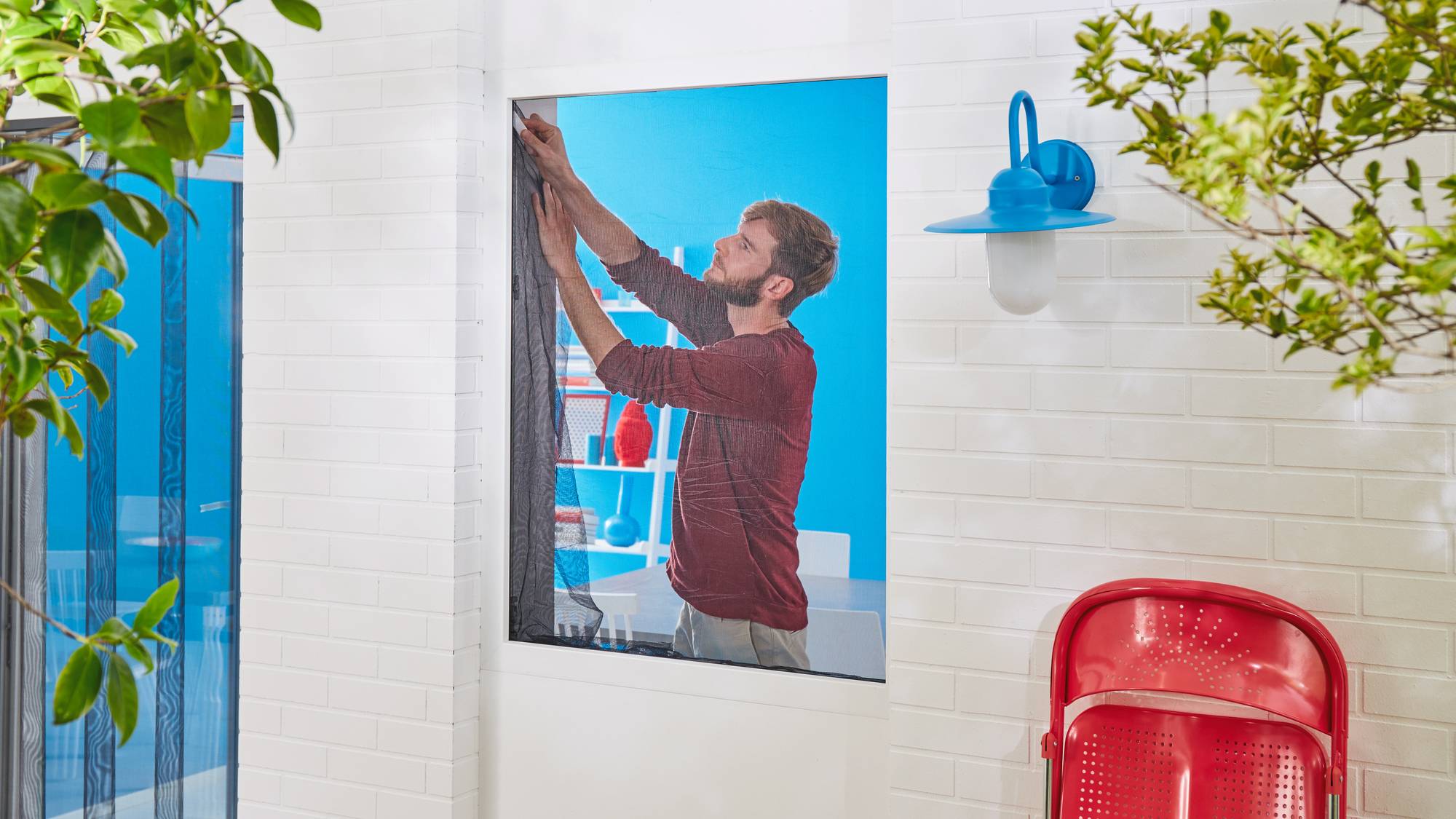 Un homme portant un pull rouge accroche un rideau. Au fond, un mur bleu vif, une chaise rouge et une lampe à l'extérieur.