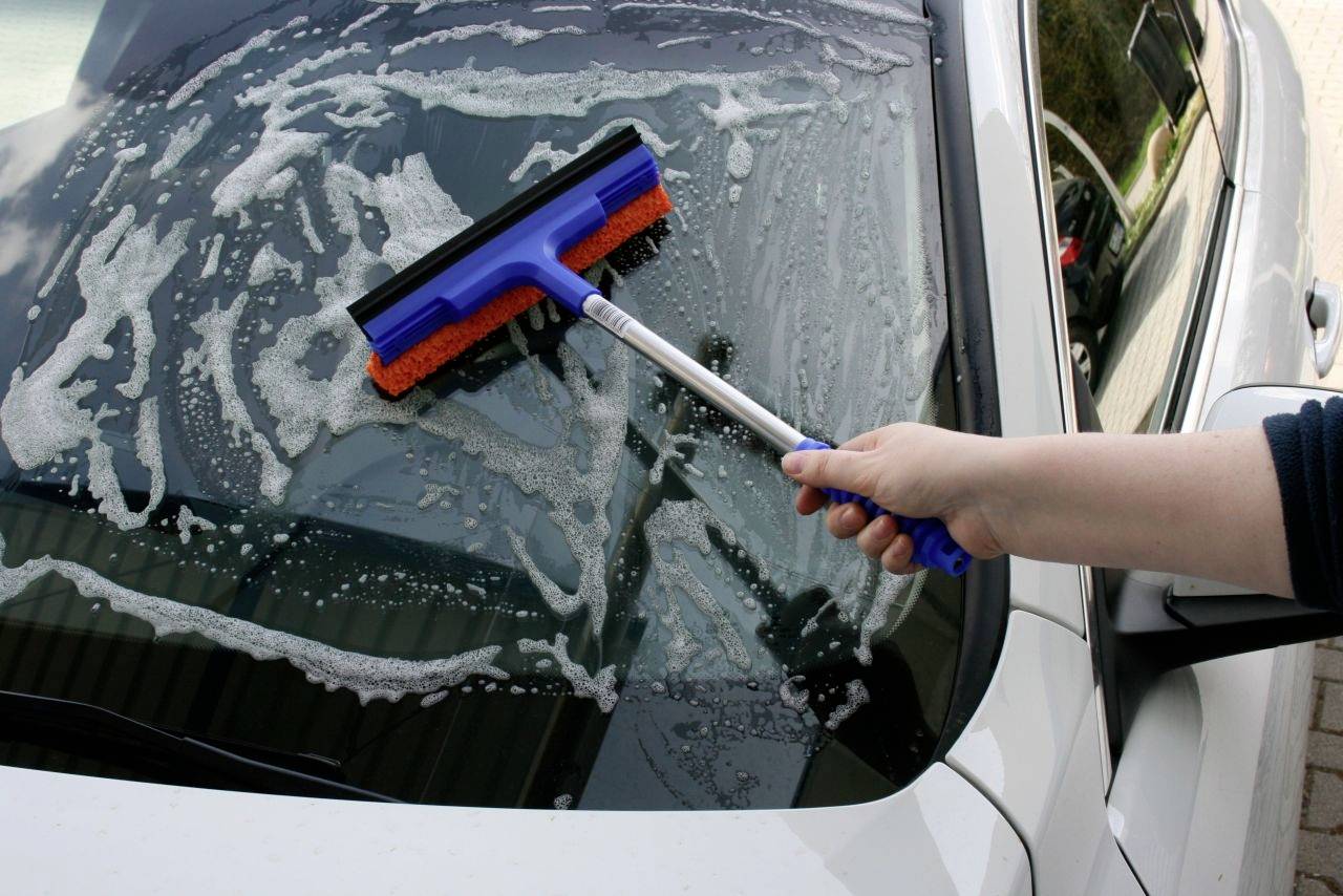 Une personne nettoie le pare-brise d'une voiture avec un raclette bleu. De l'eau savonneuse est visible sur la vitre.