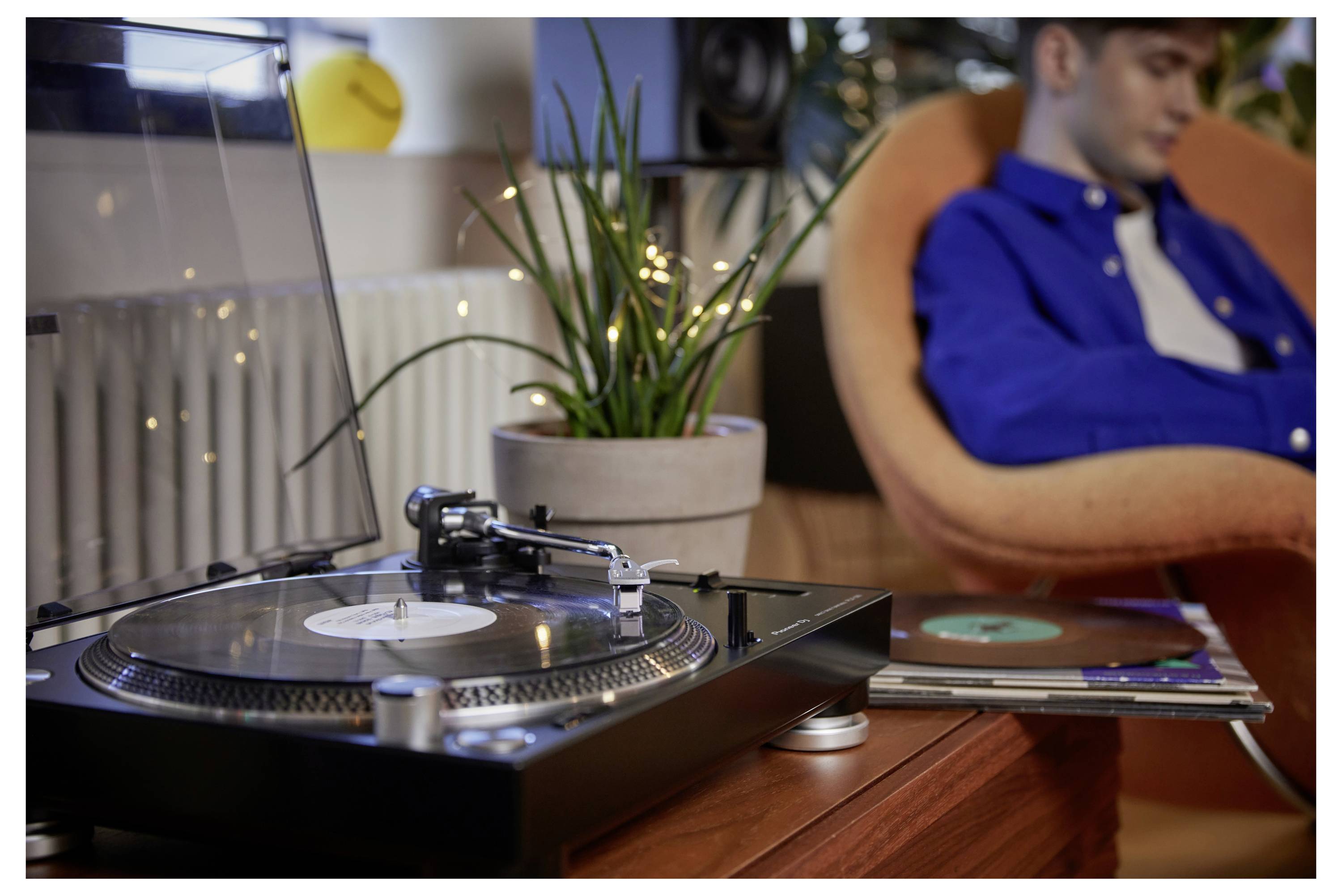 Une platine vinyle jouant un disque vinyle sur une table, avec une personne assise floue dans un fauteuil orange en arrière-plan.