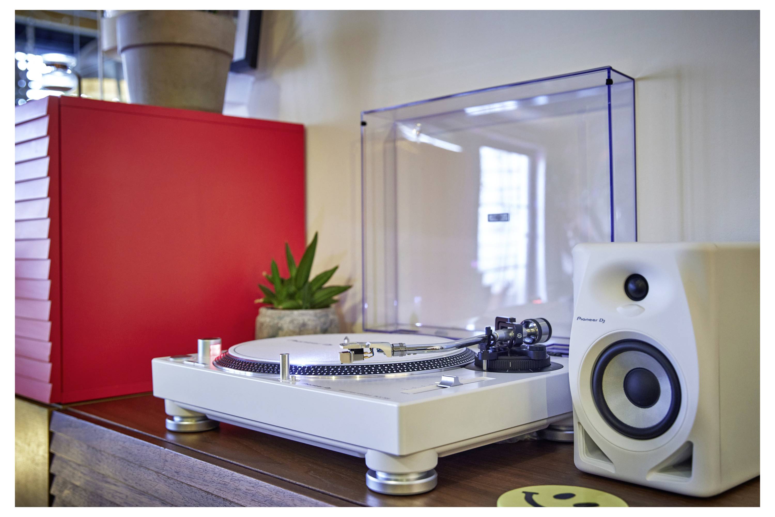 Platine vinyle blanche avec couvercle transparent anti-poussière à côté d'un haut-parleur blanc et d'un meuble rouge, avec une petite plante en pot sur le dessus du tourne-disque.
