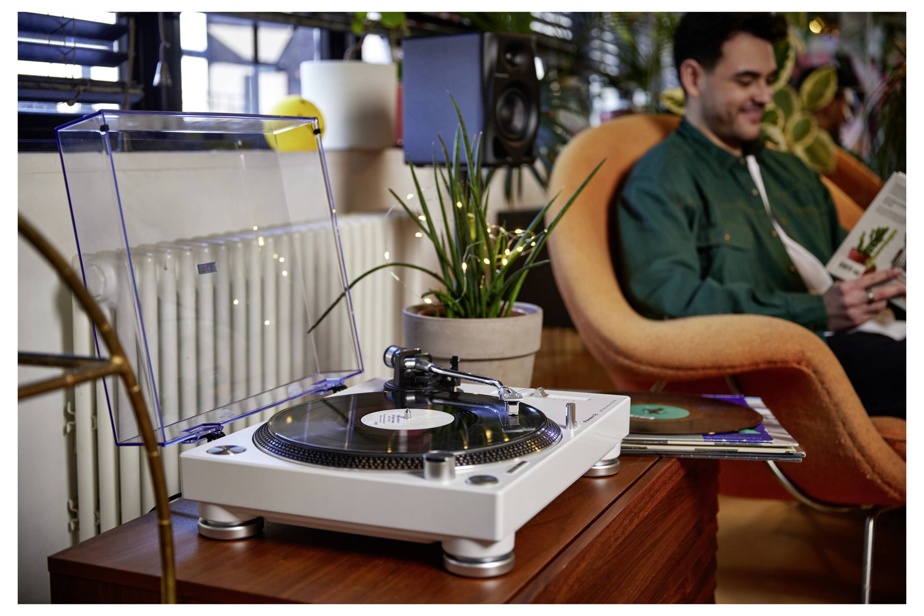 Un tourne-disque joue un disque vinyle au premier plan tandis qu'un homme est assis en train de lire dans un fauteuil en arrière-plan.