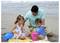 Un homme s'agenouille sur une plage avec deux enfants qui jouent dans le sable en utilisant des seaux et des pelles colorés près de l'océan.