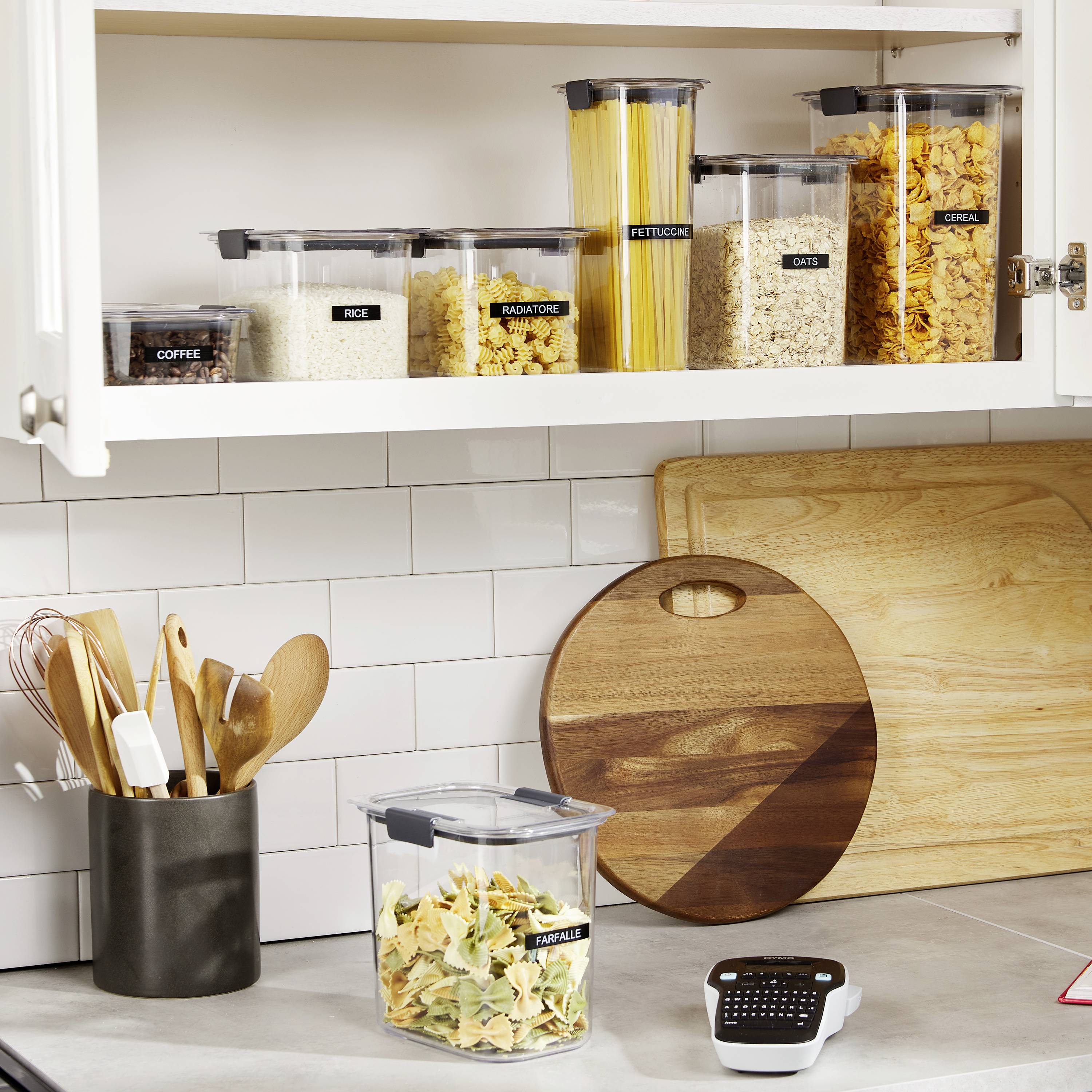 Un placard de cuisine bien rangé présente des aliments dans des contenants étiquetés : riz, pâtes, corn-flakes. Un étiqueteuse est posée sur le comptoir.