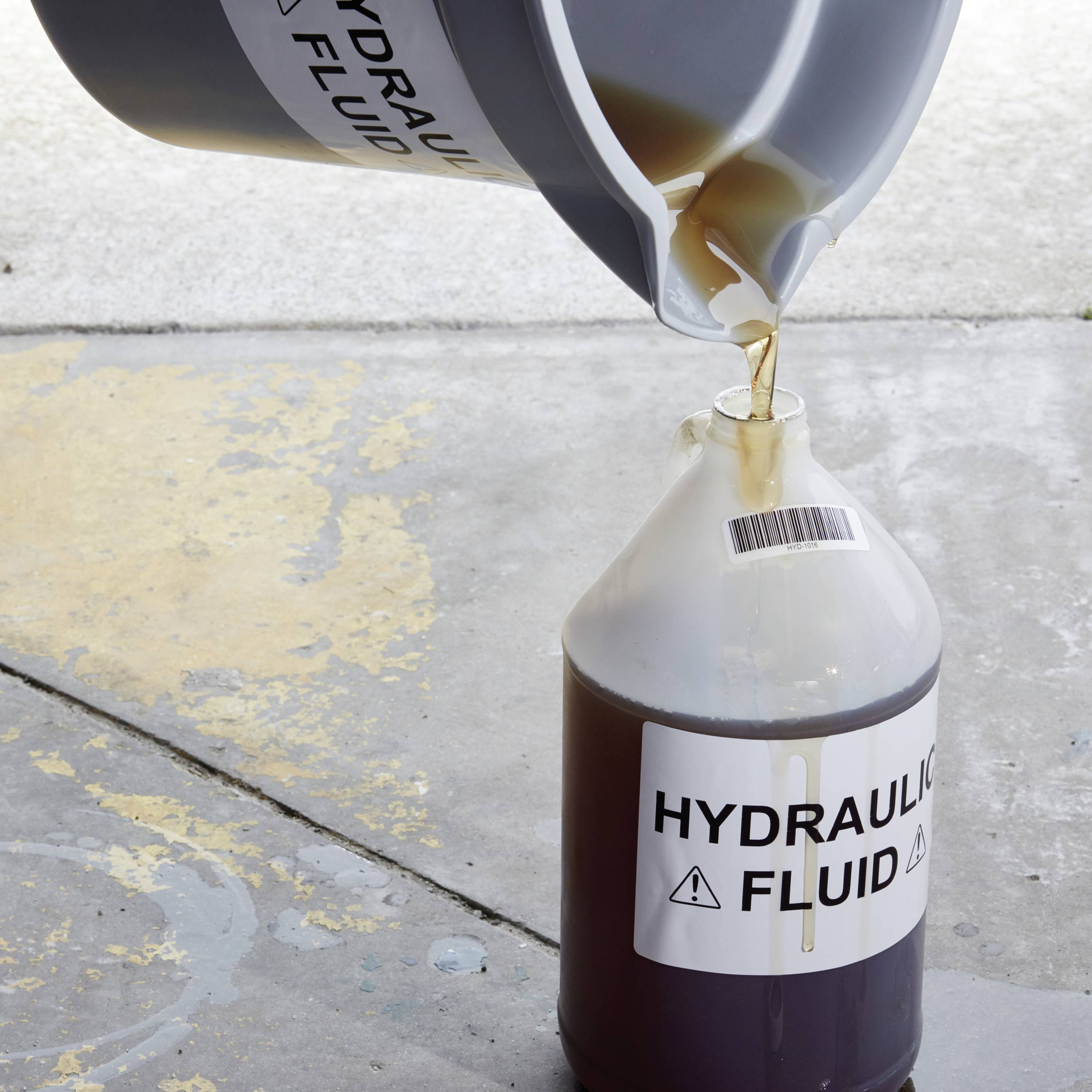 Un bidon de liquide hydraulique est transvasé dans un autre récipient. Un symbole d'avertissement de prudence est visible sur l'étiquette.