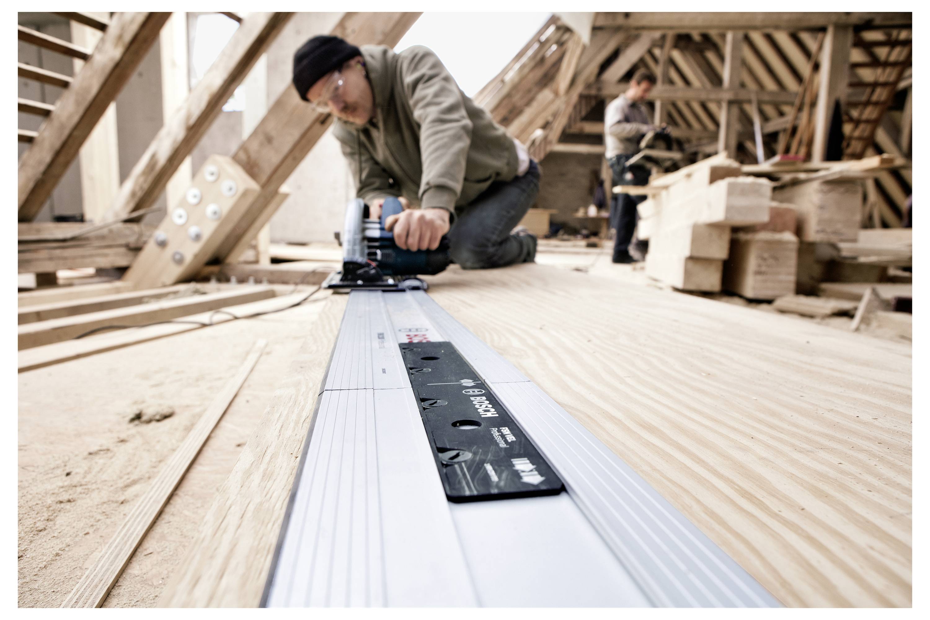 Un artisan utilise un outil électrique pour le travail du bois sur un chantier de construction. Un autre ouvrier travaille en arrière-plan.