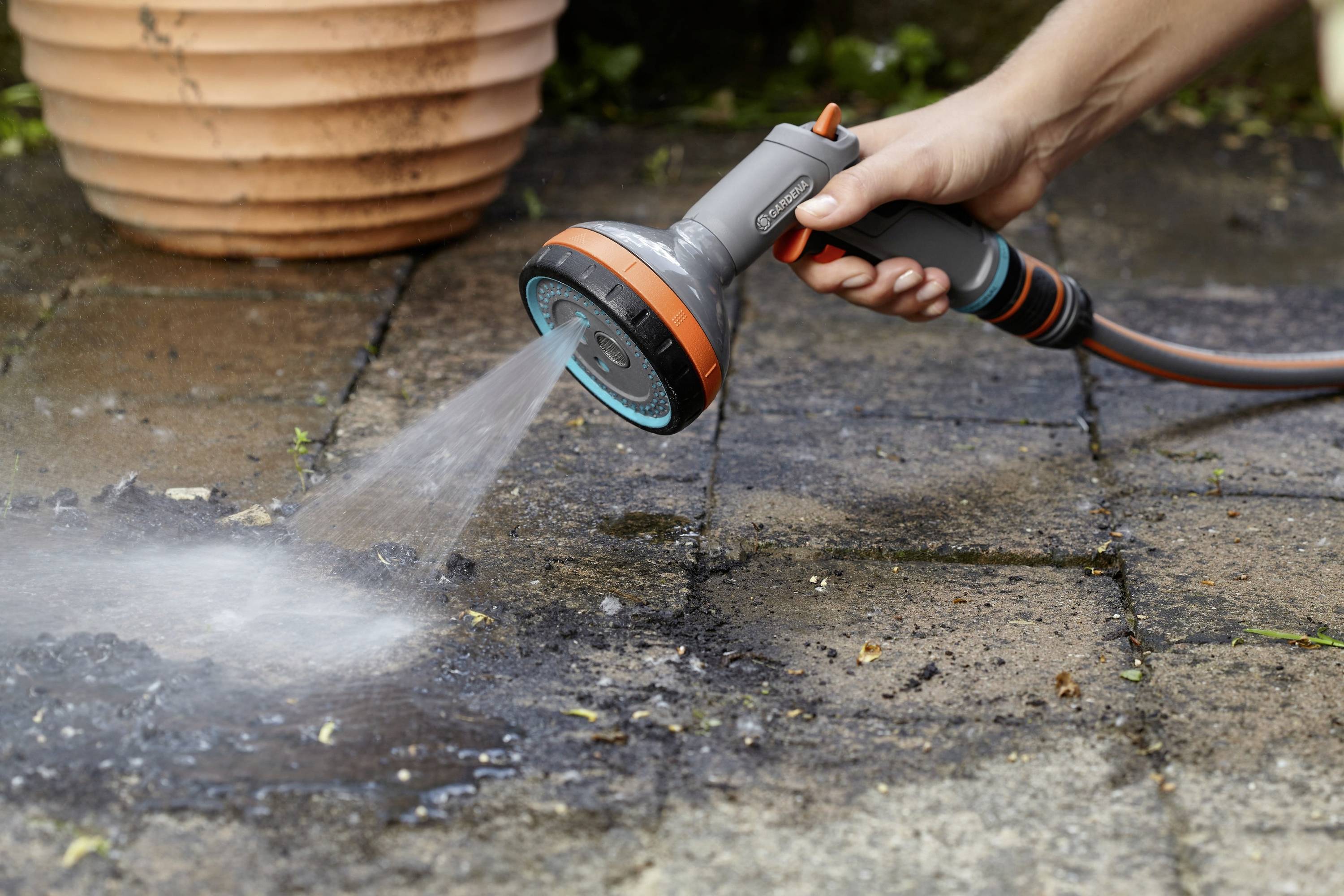 Une main tient un jet de lance d'arrosage de jardin et arrose une surface de terrasse pavée humide à côté d'un pot de fleurs.