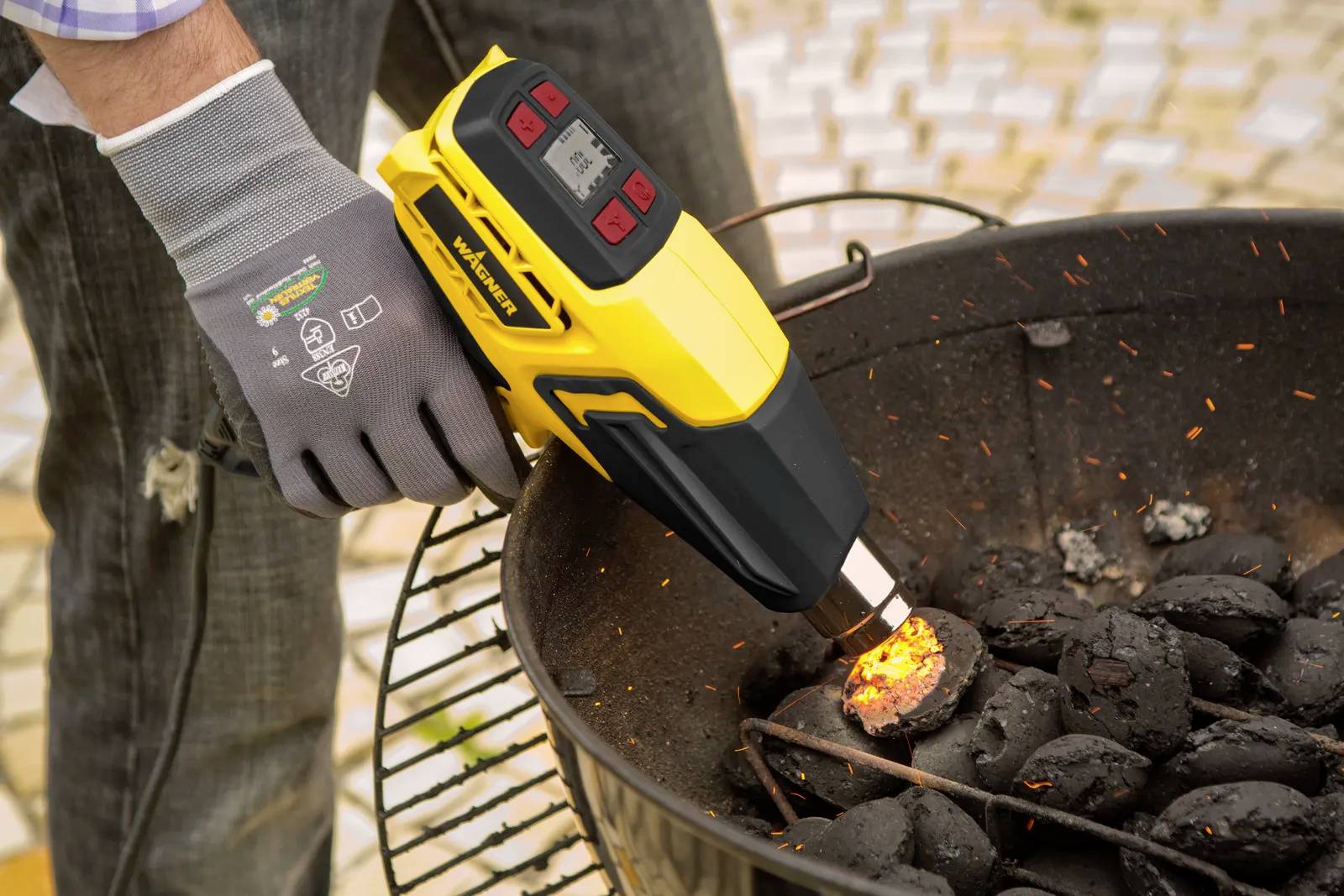 Une personne allume des briquettes de charbon dans un grill à l'aide d'un souffleur d'air chaud électrique. Les briquettes commencent à brûler.