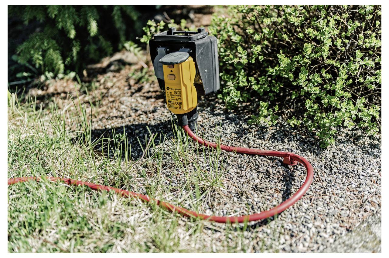 Boîtier de prise électrique extérieure jaune avec un câble rouge branché, posé sur du gravier à côté de l'herbe et des arbustes.