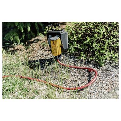 Boîtier de prise électrique extérieure jaune avec un câble rouge branché, posé sur du gravier à côté de l'herbe et des arbustes.