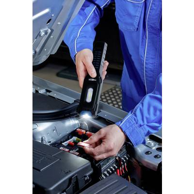 Une personne en vêtements de travail bleus vérifie les fusibles dans un moteur de voiture ouvert à l'aide d'une lampe de poche.