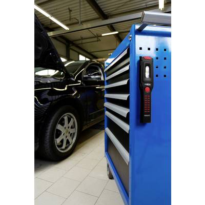 Une voiture noire, capot ouvert, est stationnée dans un atelier. Au premier plan, on aperçoit un chariot à outils bleu équipé d'une lampe.