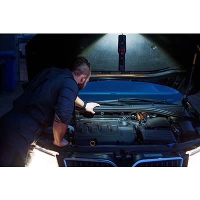 Un mécanicien inspecte le compartiment moteur d'une voiture sous un capot ouvert. Une lampe éclaire la zone.