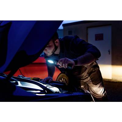 Une personne examine le compartiment moteur d'une voiture à l'aide d'une lampe de poche au crépuscule.