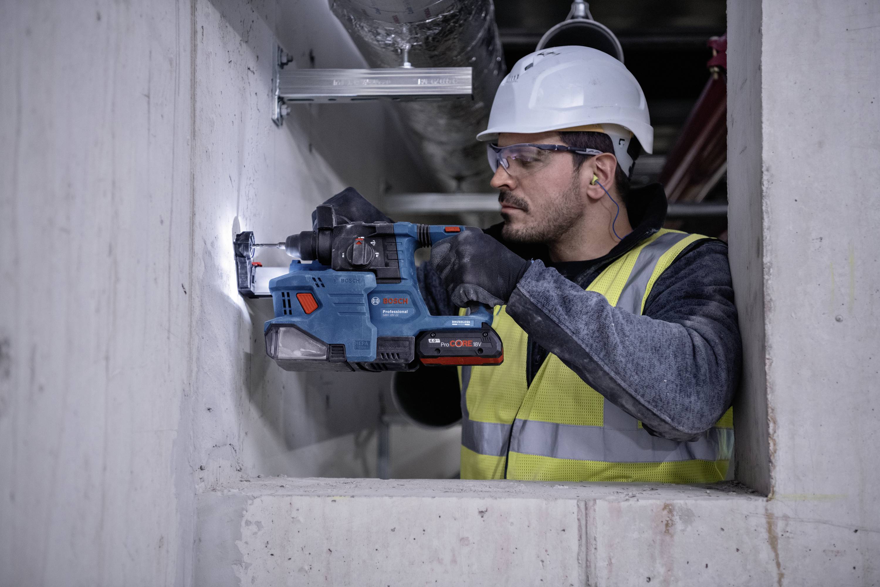 Un ouvrier porte un casque et un gilet de sécurité et fore un trou dans un mur en béton à l'aide d'une perceuse électrique.