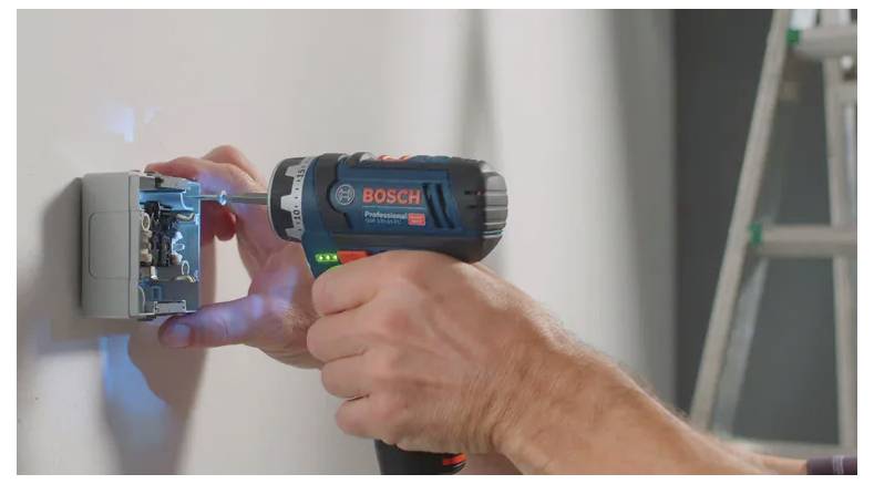 Une personne utilise une perceuse sans fil Bosch pour travailler sur un boîtier électrique monté sur un mur. Un escabeau est visible en arrière-plan.