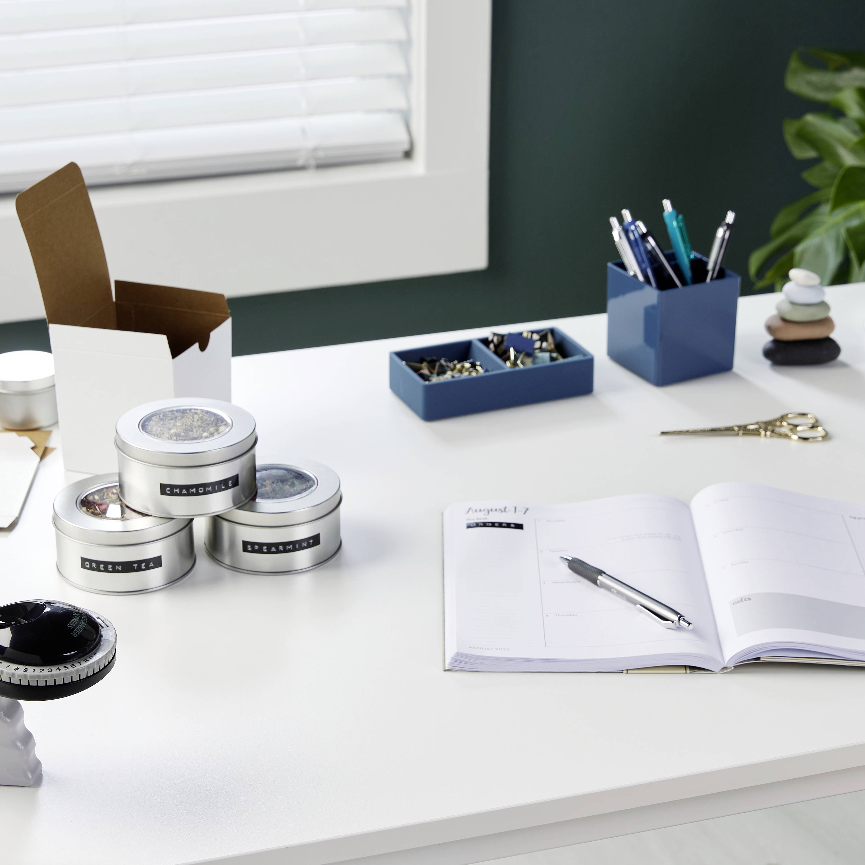 Un bureau organisé avec un agenda ouvert, des stylos, des boîtes et un cactus devant une fenêtre à stores.