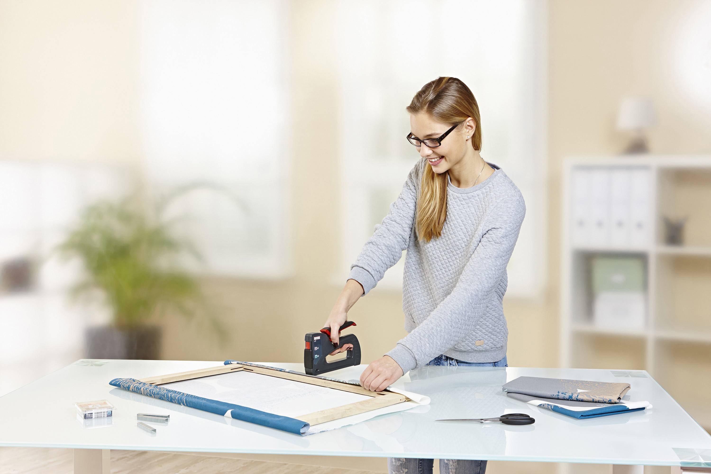 Une femme portant un pull gris clair utilise une agrafeuse pour fixer un cadre sur une table. Un espace de bureau est visible en arrière-plan.