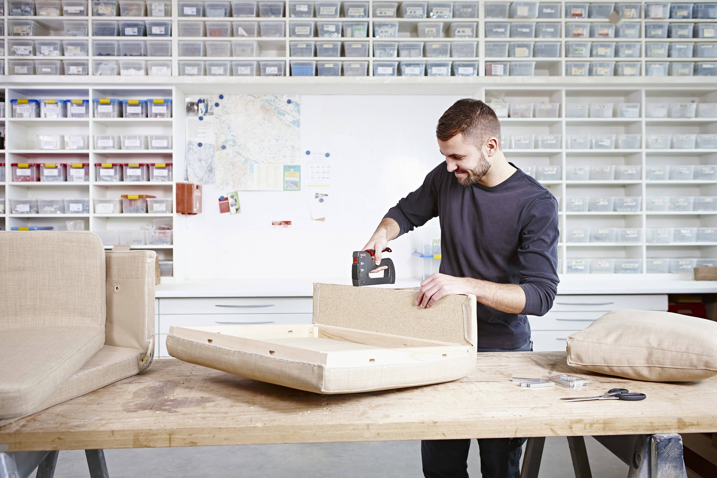 Un homme rembourre un meuble dans un atelier avec des étagères remplies de matériaux en arrière-plan.