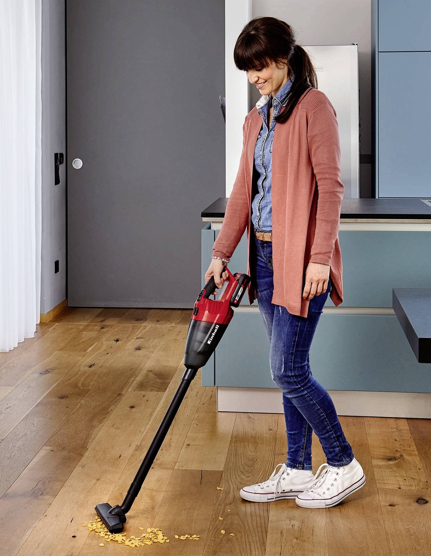 Une femme aspire des miettes du plancher en bois dans une cuisine moderne à l'aide d'un aspirateur.