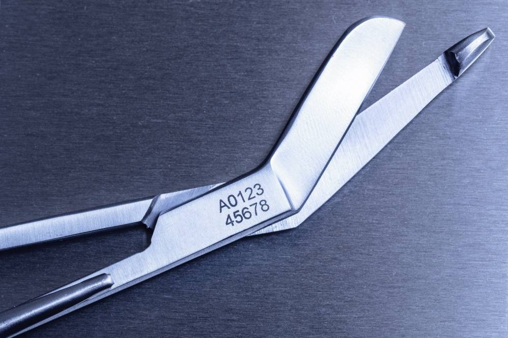 Un instrument chirurgical métallique portant la gravure 'A0123 45678', posé ouvert sur un fond sombre.